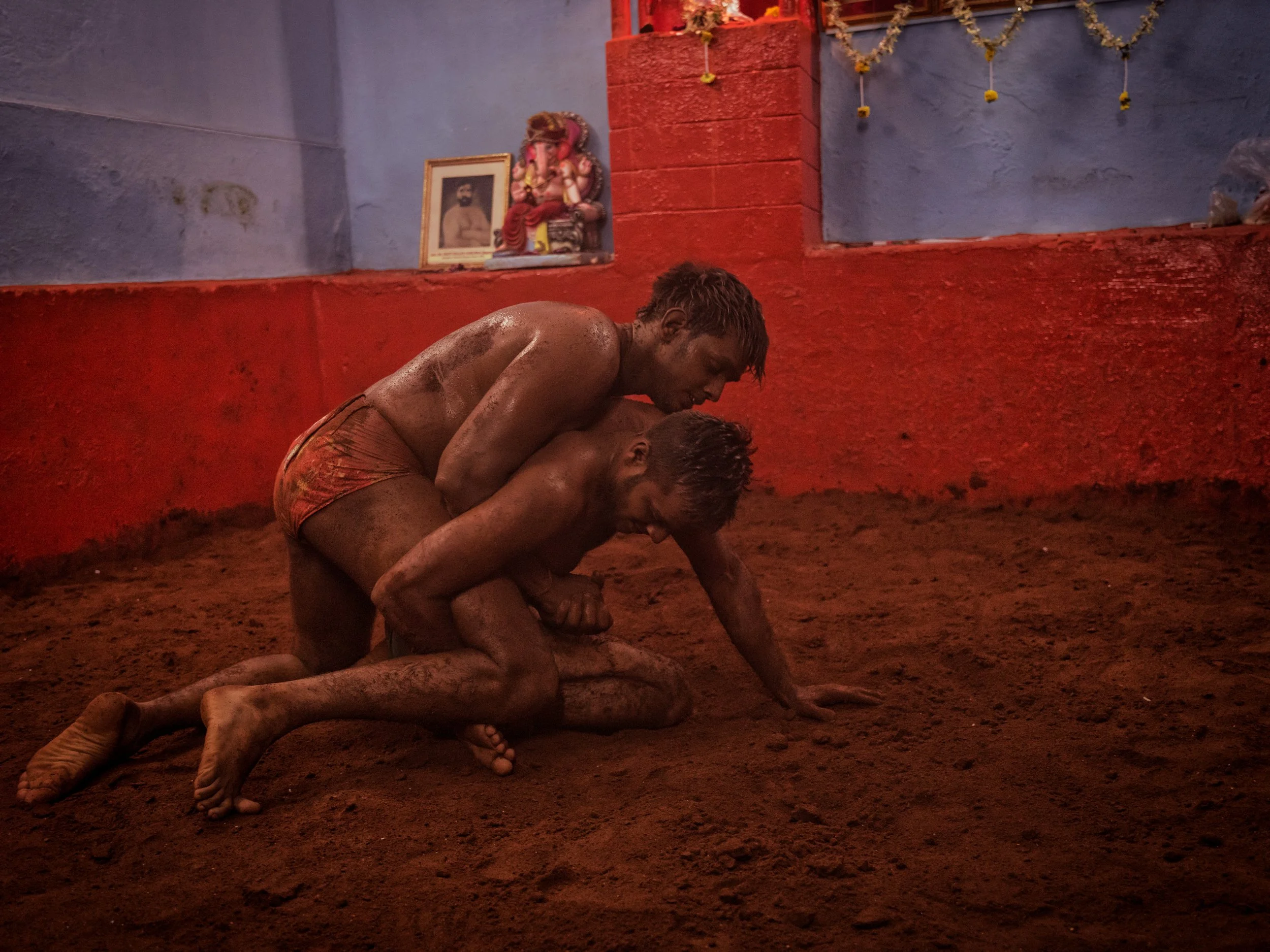 Two traditional Indian wrestlers grapple on the red mud floor of a kushti akhara near a red shrine, Ganesh idol, framed portrait, and flower garlands.