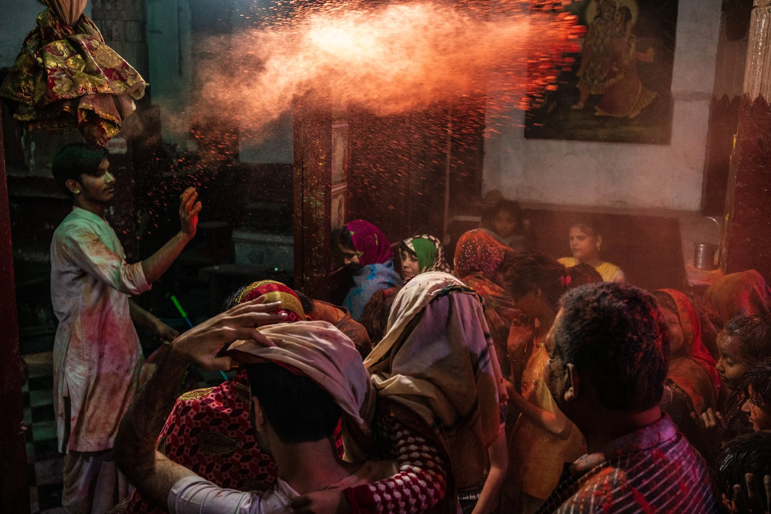 Young man throws orange Holi powder over a crowd of women and children inside a temple, creating a glowing cloud of gulal.