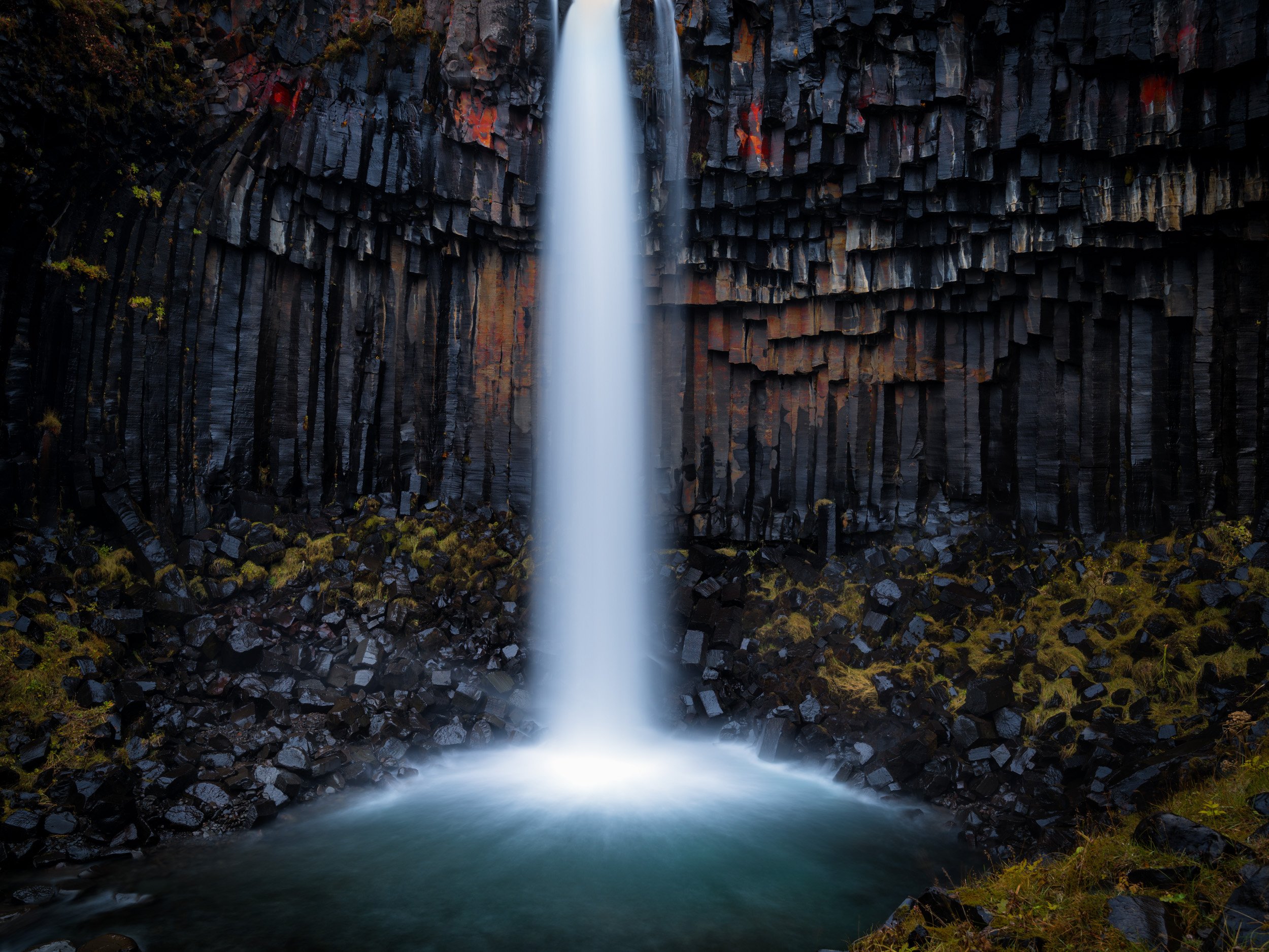 Tall Iceland waterfall falling into a misty turquoise pool, framed by dark basalt columns, volcanic rock, and mossy green stones.
