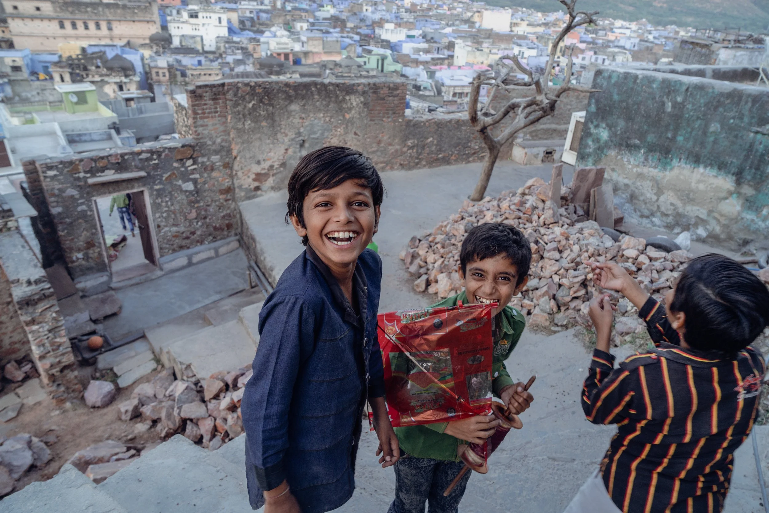 Smiling children fly a red kite from a rooftop overlooking a dense cityscape and old stone walls.