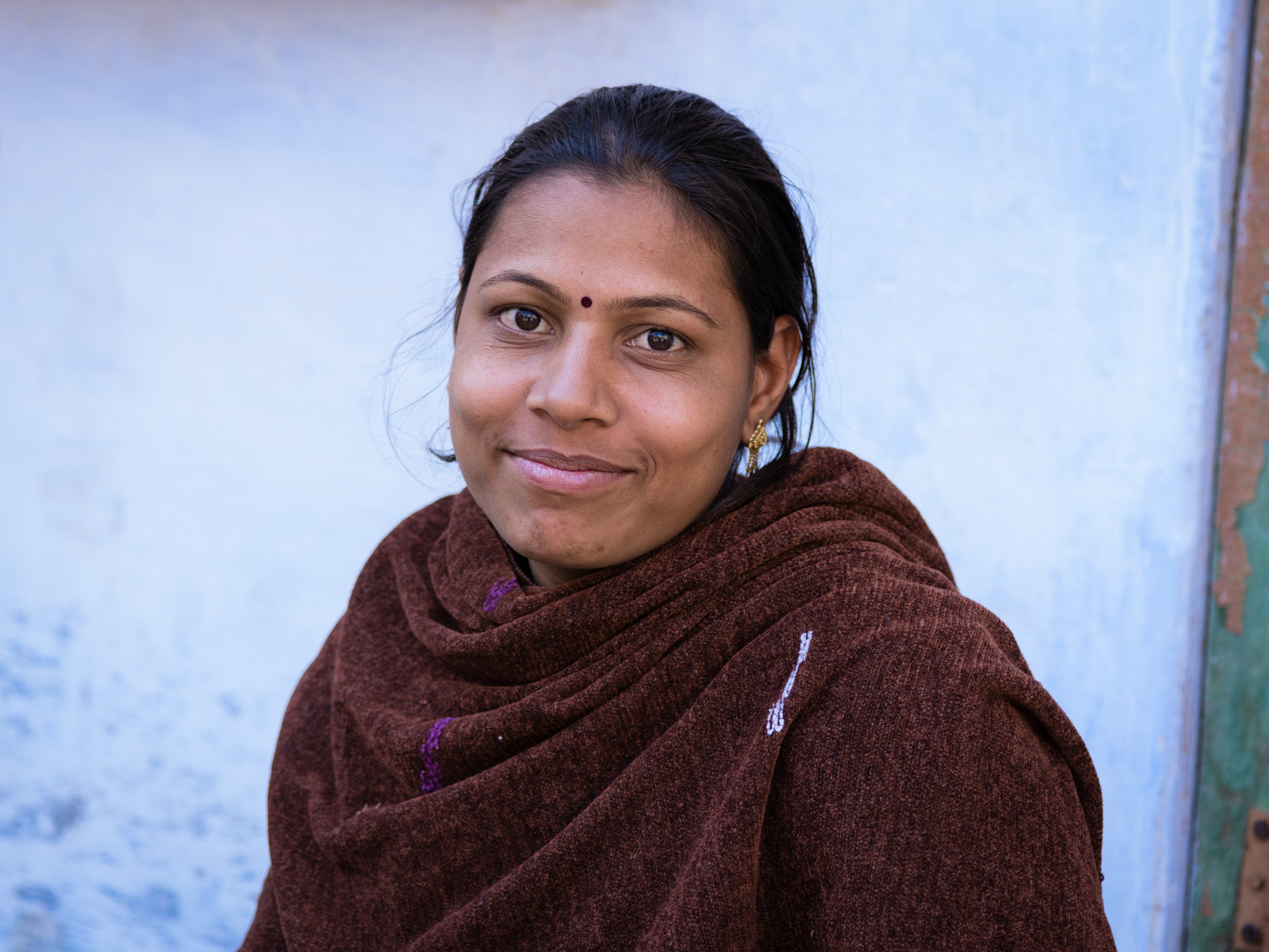 Young Indian woman with a small bindi and brown shawl smiles softly against a pale blue wall in a natural light travel portrait.