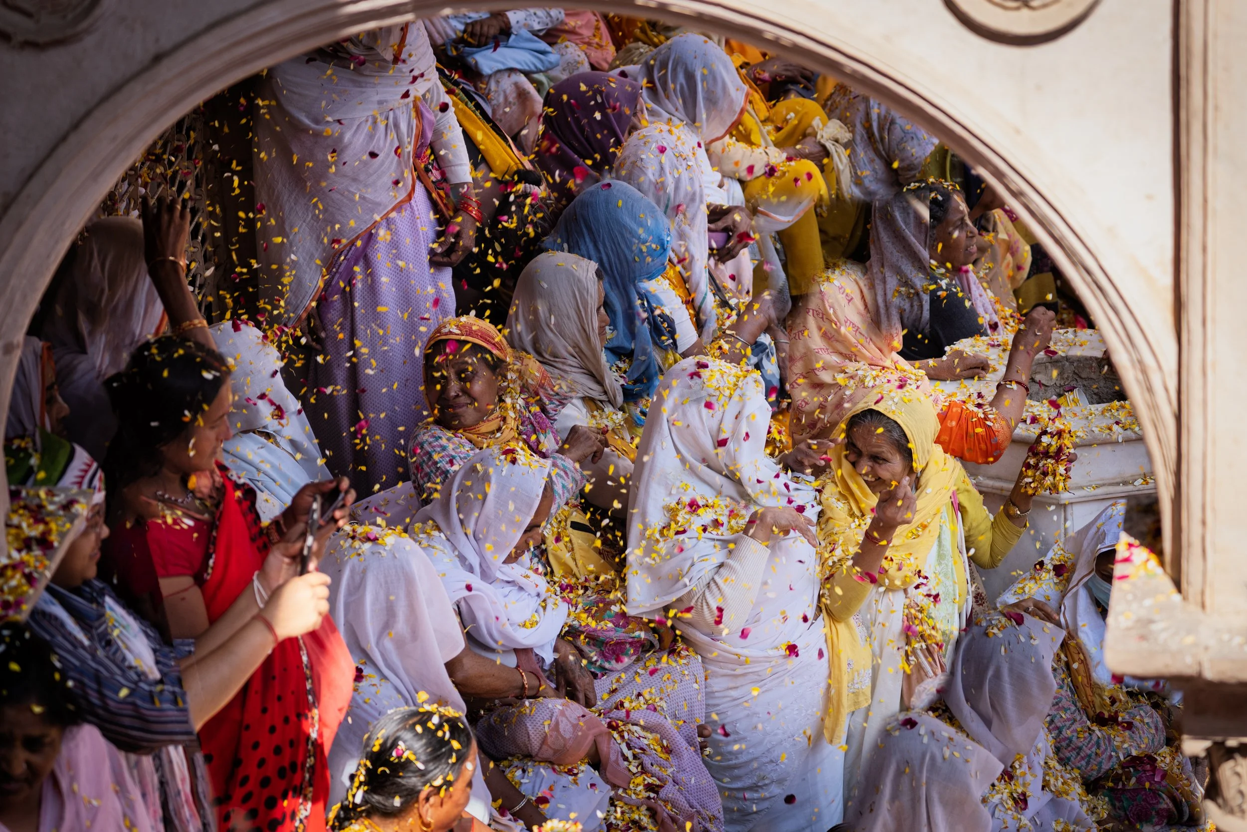 Women in white and yellow sarees sit beneath an arched temple balcony during a Holi flower festival, covered in yellow marigold and rose petals during a devotional Hindu celebration.