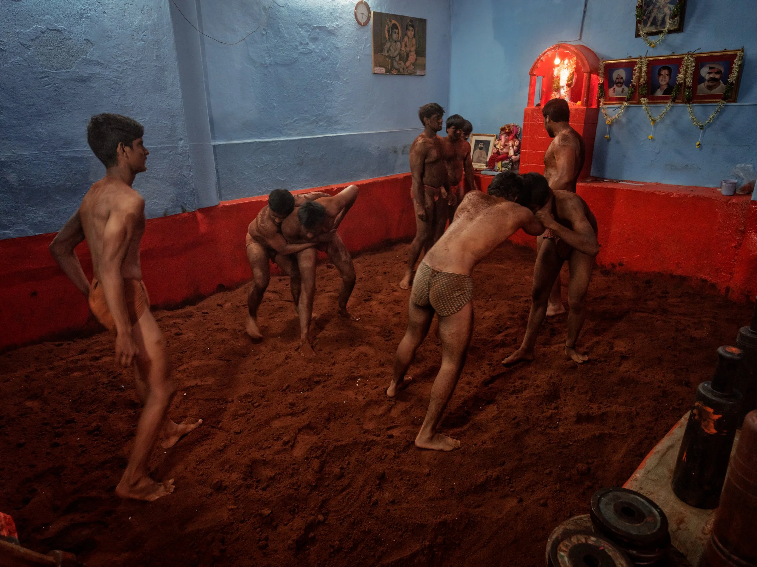 Wide view of a traditional Indian kushti akhara where shirtless wrestlers practice grappling and conditioning on a red mud floor beneath blue walls, religious portraits, and a glowing shrine.