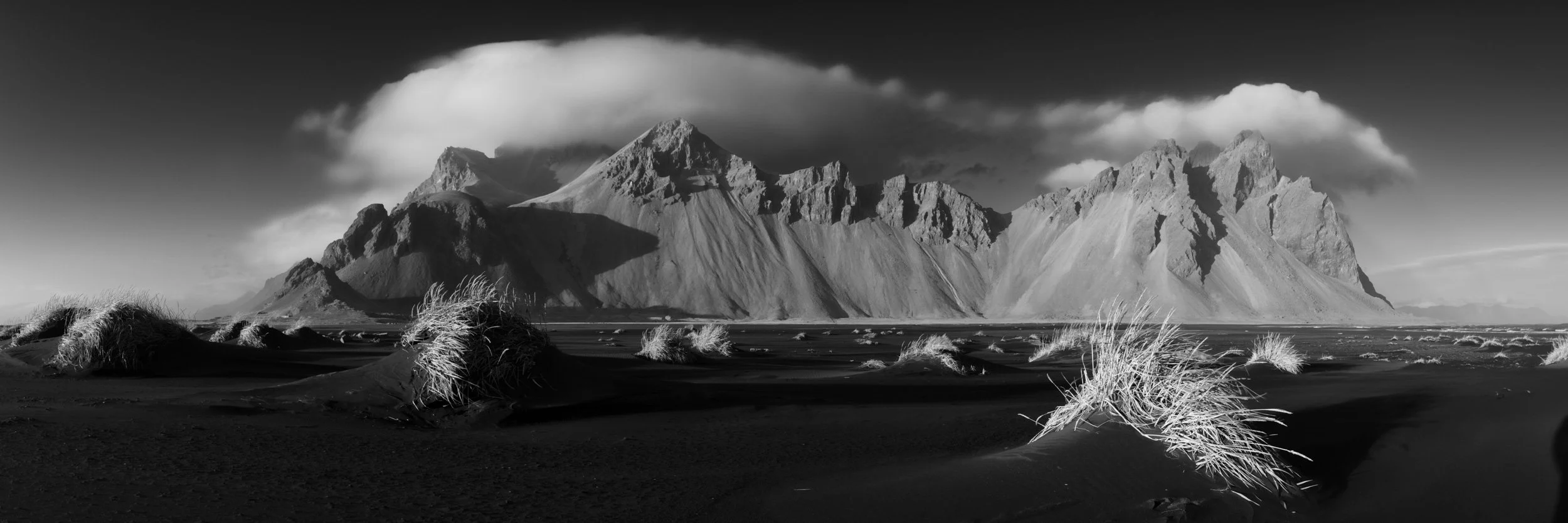 Black and white Iceland mountain landscape with black sand, windblown grass, and dramatic cloud-covered peaks.