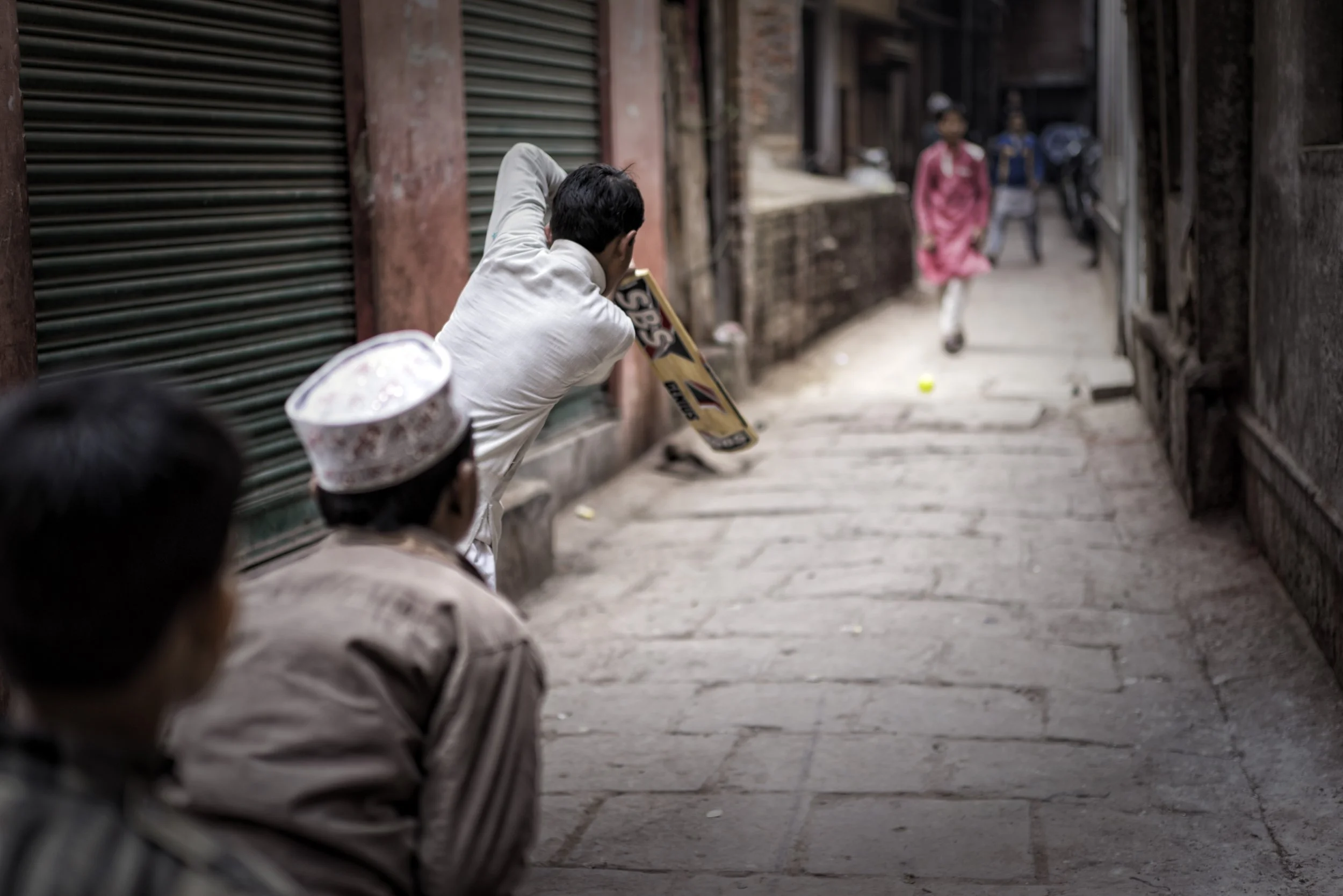 Children play street cricket in a narrow alley, with a boy swinging a bat toward a tennis ball on the pavement.