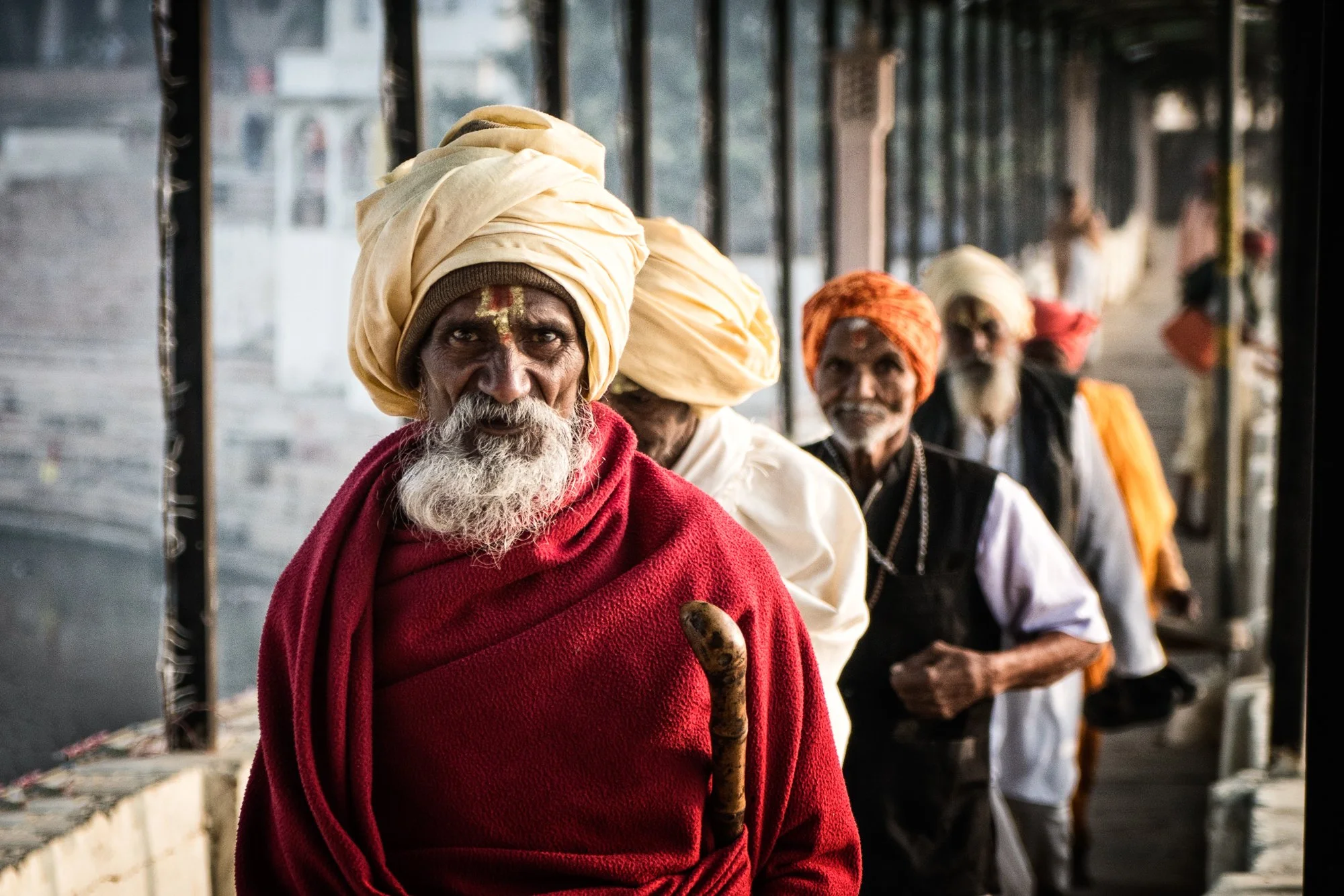 Elderly Sikh men in cream turbans sit in a row, with one man wrapped in a red shawl holding a walking stick.