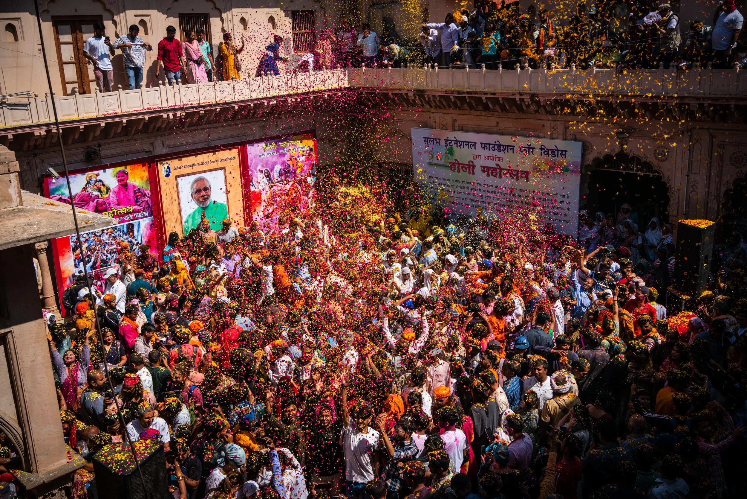 Large crowd celebrates Holi Mahotsav in an Indian temple courtyard as colorful flower petals rain from balconies, with visible signs reading “Sulabh International Social Service Organisation” and “होली महोत्सव.”