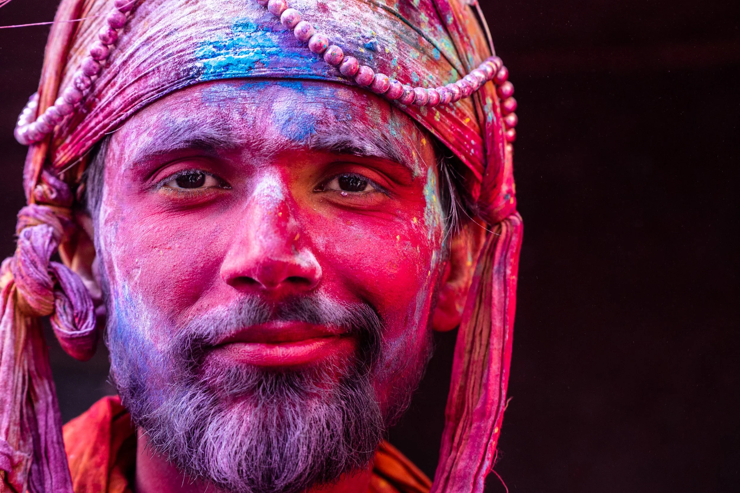 Close-up portrait of a man wearing a decorative turban, his face covered in vivid pink, blue, and red Holi festival powder.