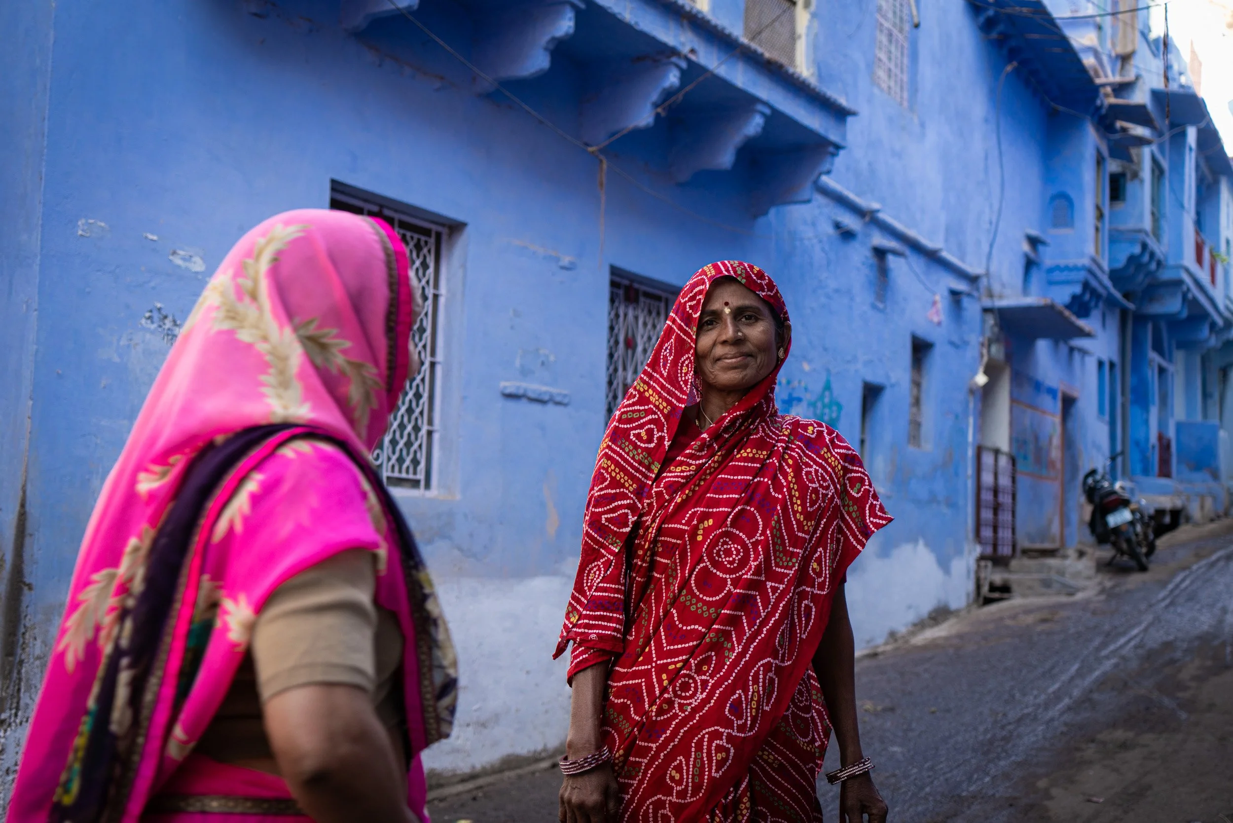 Women in bright red and pink sarees stand in a blue-painted alley, creating a colorful Indian street photography scene in Bundi, India.