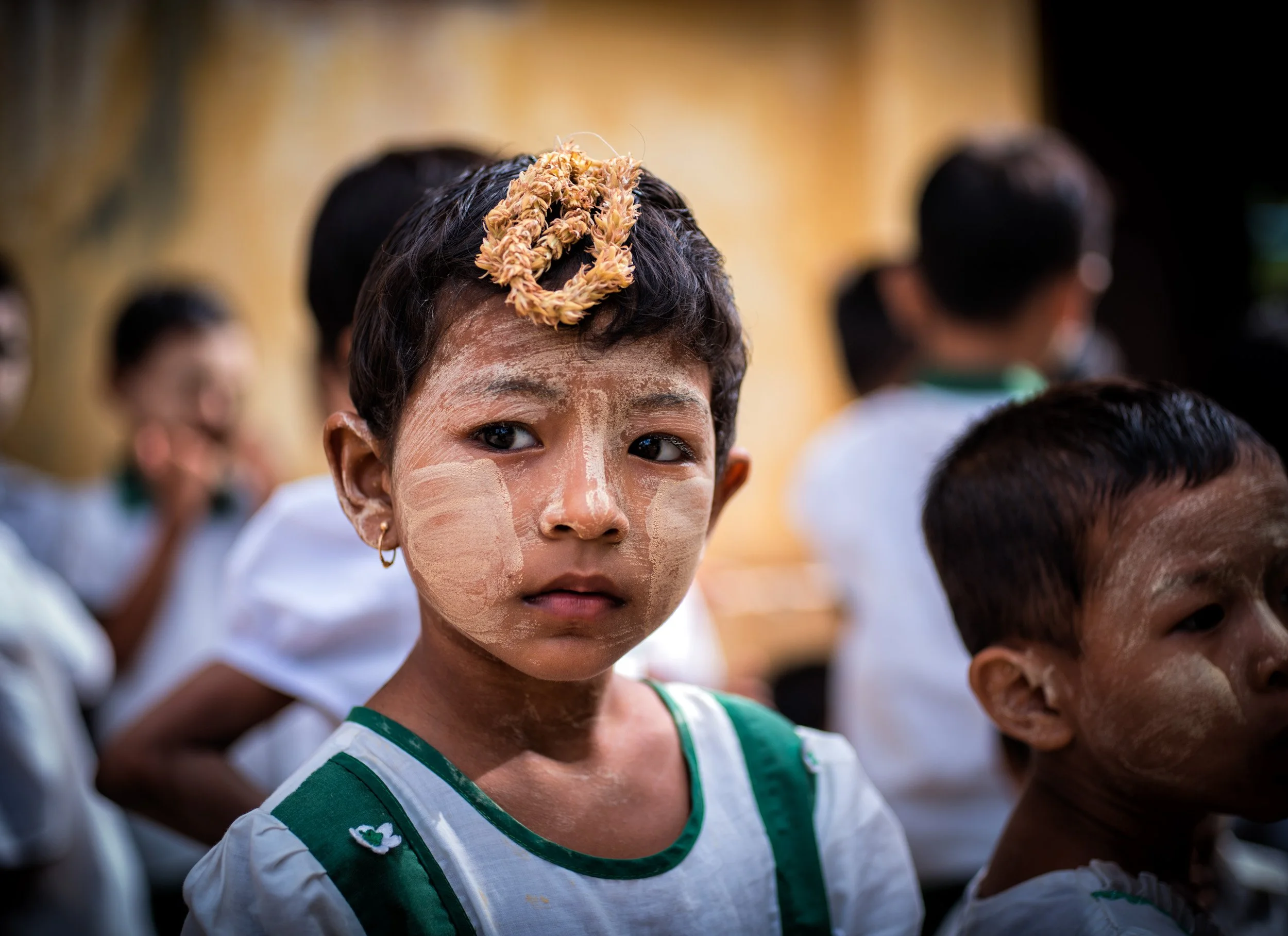Young Myanmar child with traditional face paint and a flower headpiece looks at the camera during a cultural portrait session.