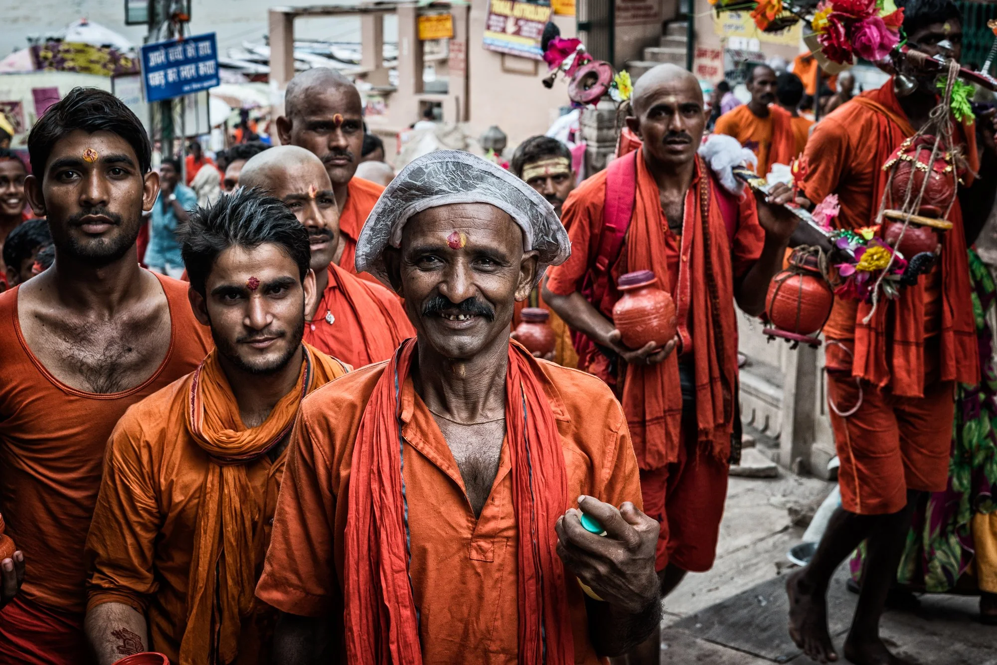 Hindu pilgrims in orange clothing gather during a religious procession, carrying water pots and decorated offerings in Varanasi, India.