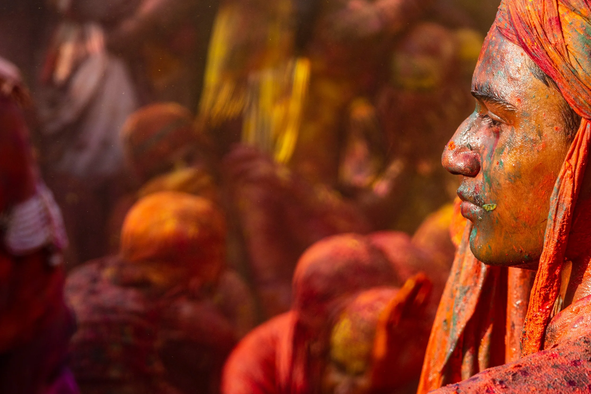 Man in an orange headscarf with blue and red Holi powder on his face watches the colorful festival crowd in warm sunlight.