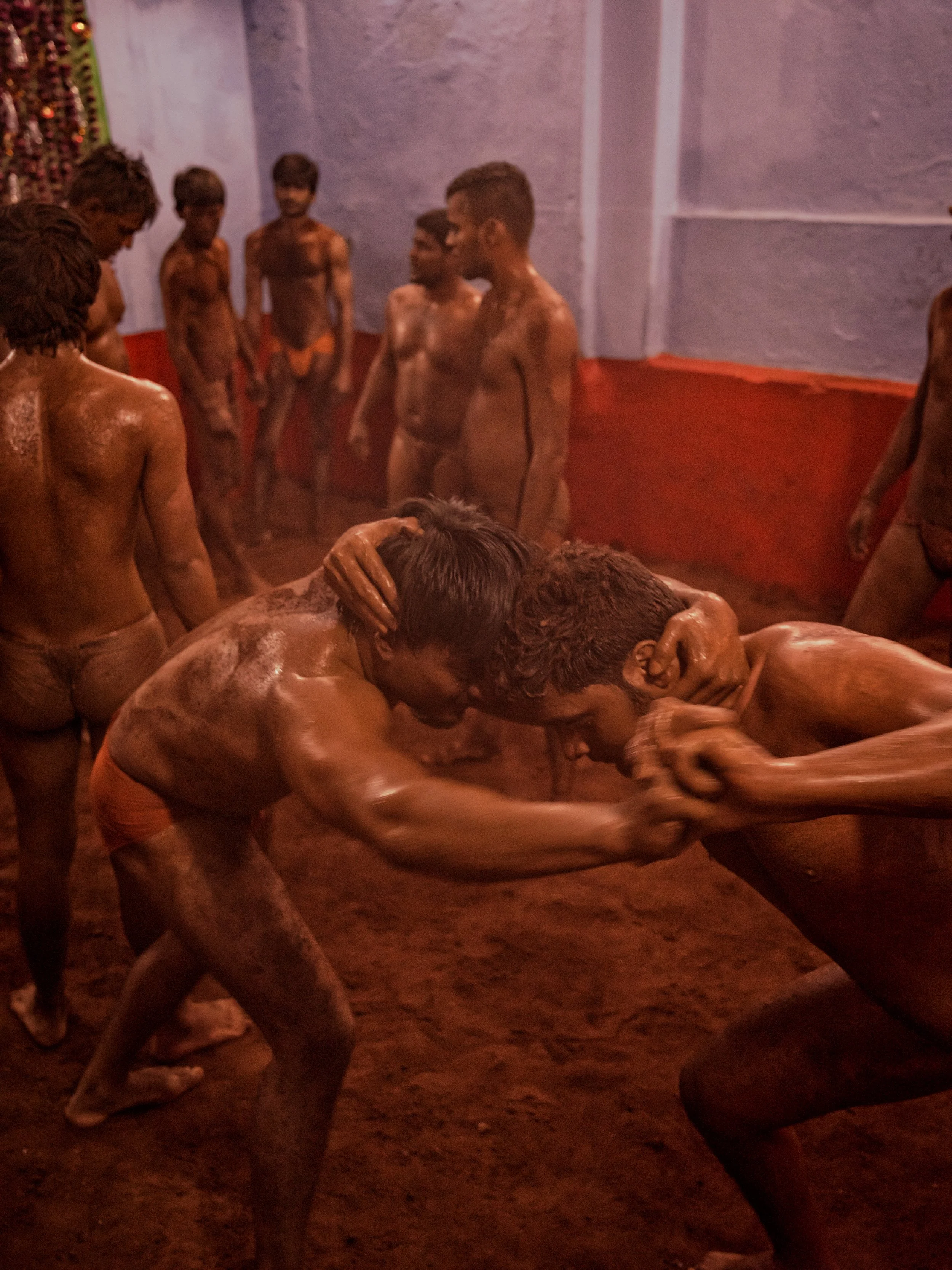 Two mud-covered wrestlers grapple in a low stance during traditional Indian kushti practice, surrounded by other trainees in a hazy red clay wrestling arena.