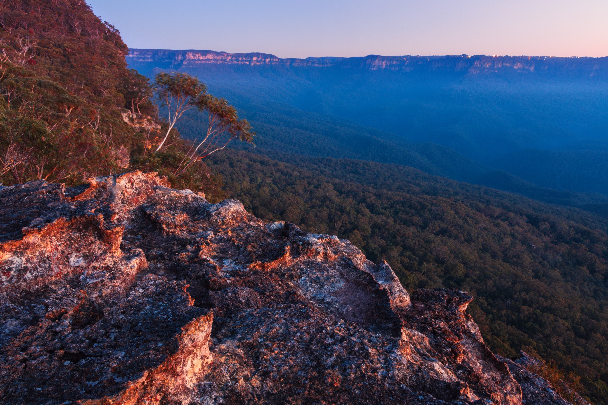 Blue Mountains cliff lookout in New South Wales, Australia, with rocky escarpment, eucalyptus forest, and distant hazy ridges.
