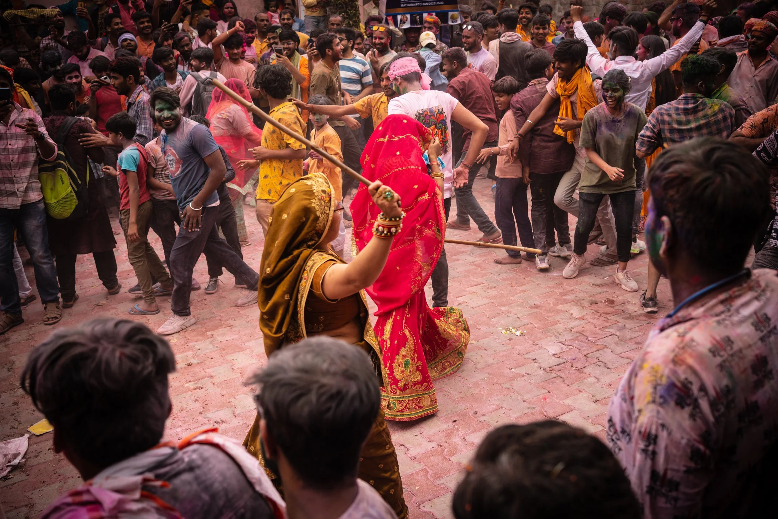 Two women in traditional red and yellow sarees raise wooden sticks during Lathmar Holi celebrations as a joyful crowd dances through clouds of colored powder.