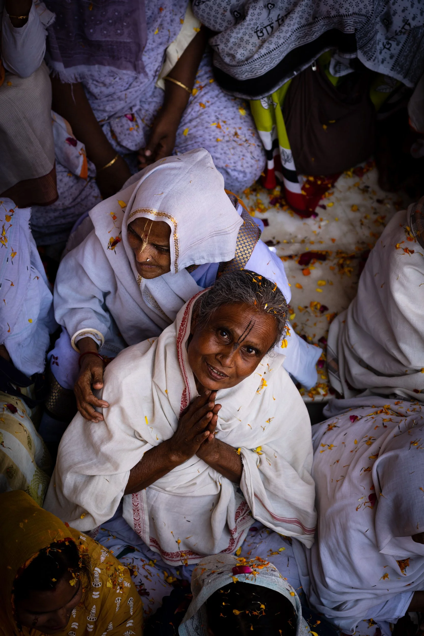 Elderly woman in a white shawl folds her hands in prayer during a Hindu Holi flower celebration, surrounded by women in white clothing and scattered yellow marigold petals.