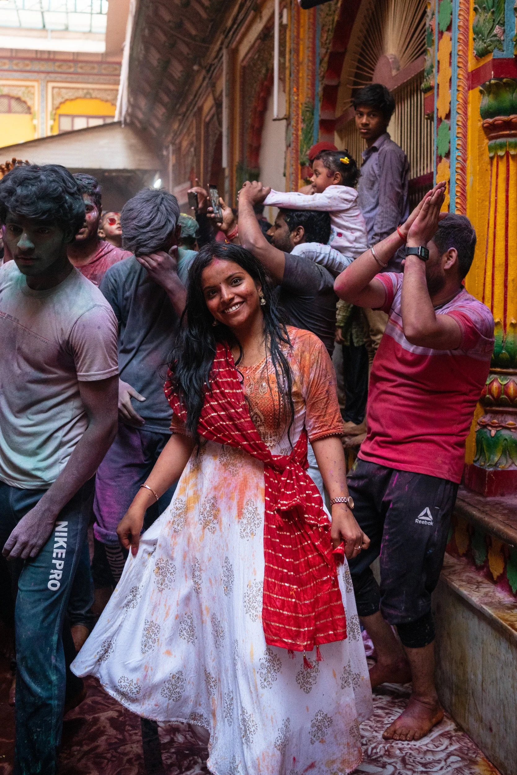 Smiling woman in a white dress and red dupatta poses in a crowded temple during Holi, with festivalgoers covered in colored powder behind her.