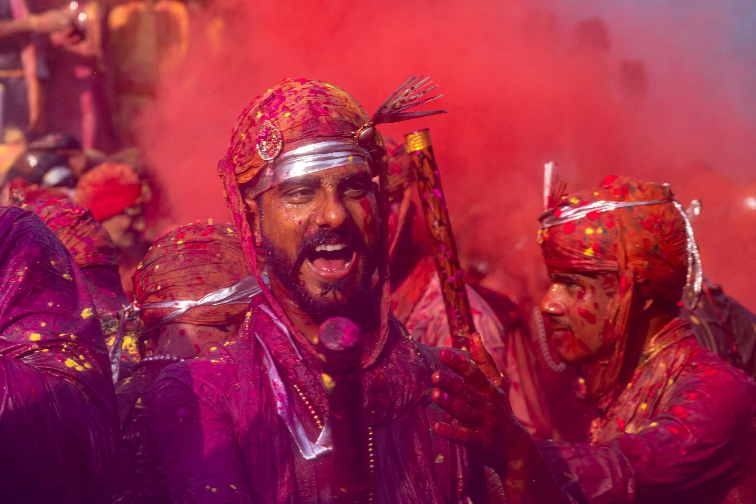 Men wearing traditional turbans celebrate Holi in a haze of red and pink powder, surrounded by vivid gulal and festive energy.