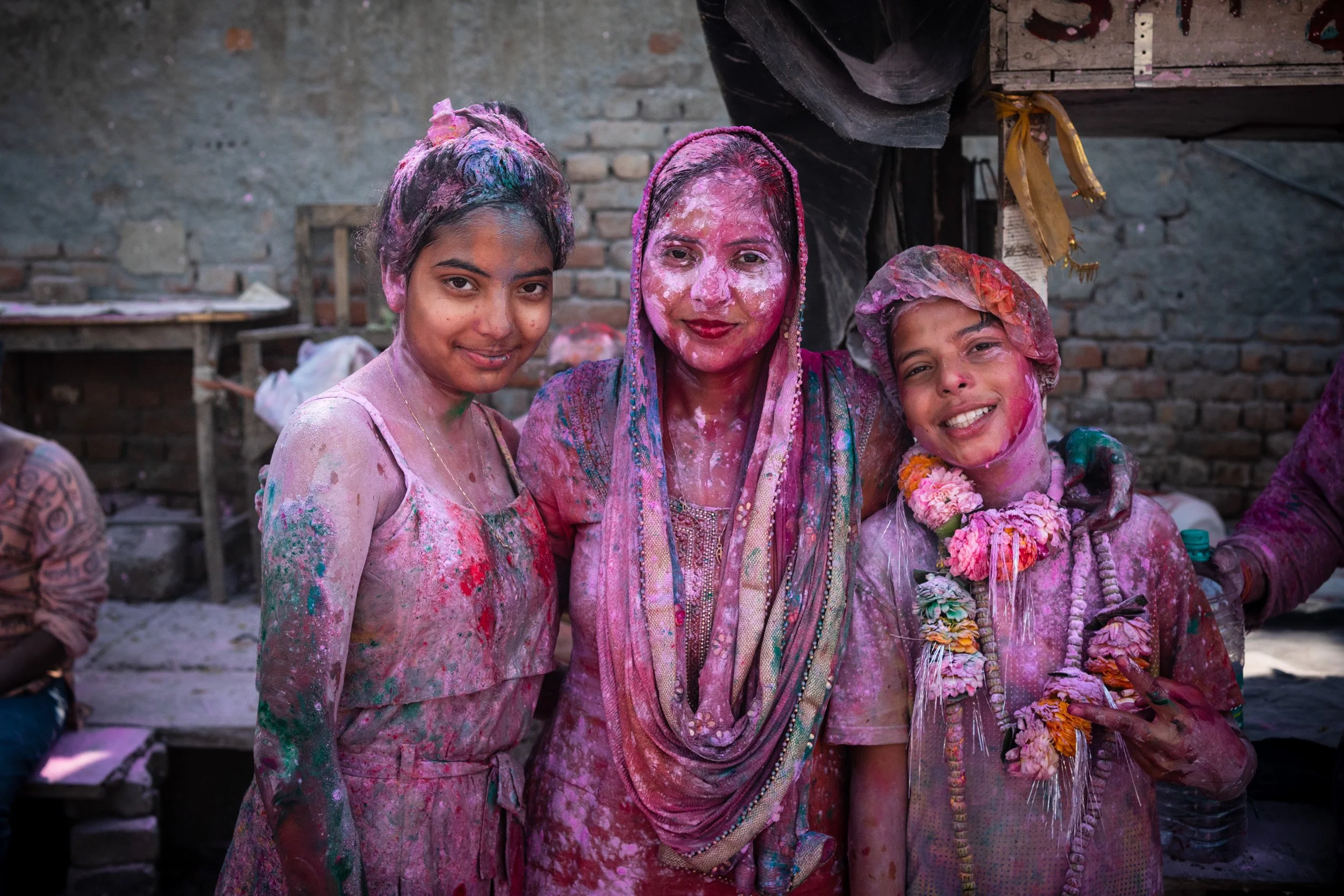 Family and friends pose after Holi celebrations, covered in pink, green, and purple gulal powder, with a smiling child wearing flower garlands.
