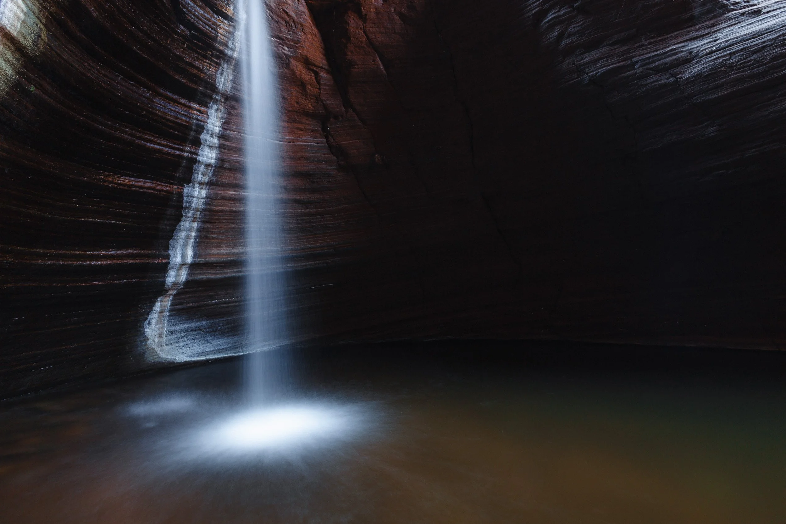 Waterfall pouring into a dark gorge pool at Karijini National Park, surrounded by layered red rock walls.