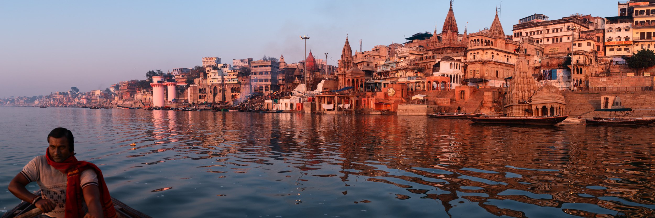 Panoramic view of Varanasi ghats along the Ganges River at sunrise, with historic riverside buildings reflected in calm water.