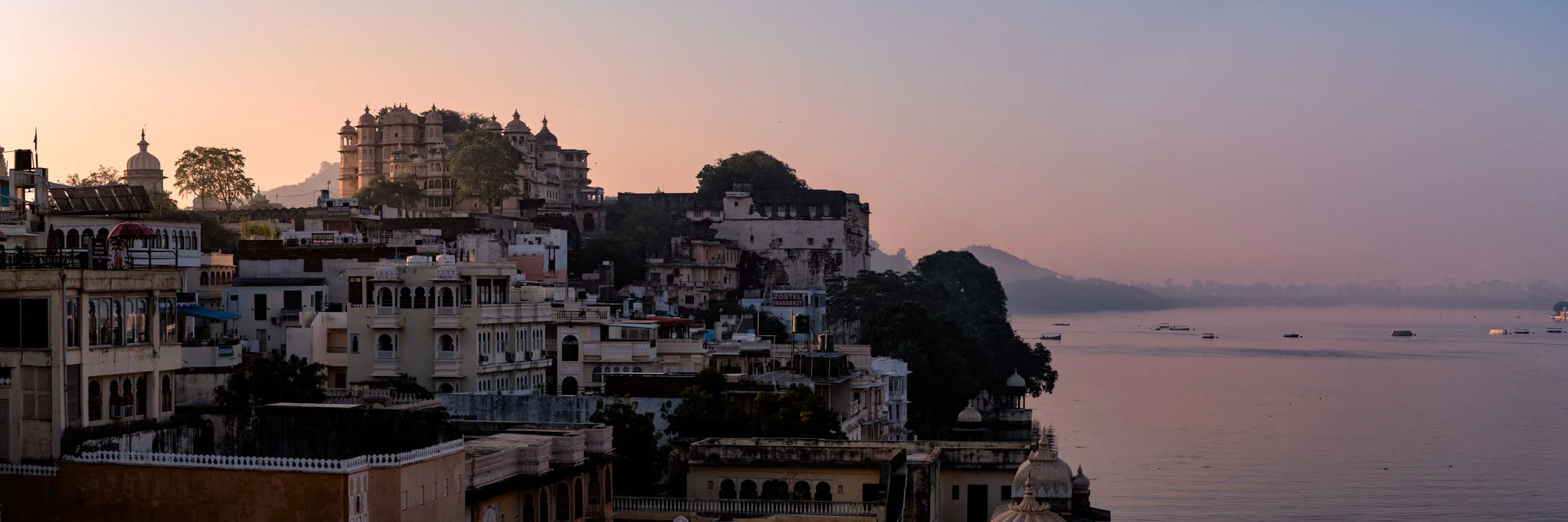 Udaipur City Palace overlooking Lake Pichola at sunset, with layered historic buildings and soft evening light.