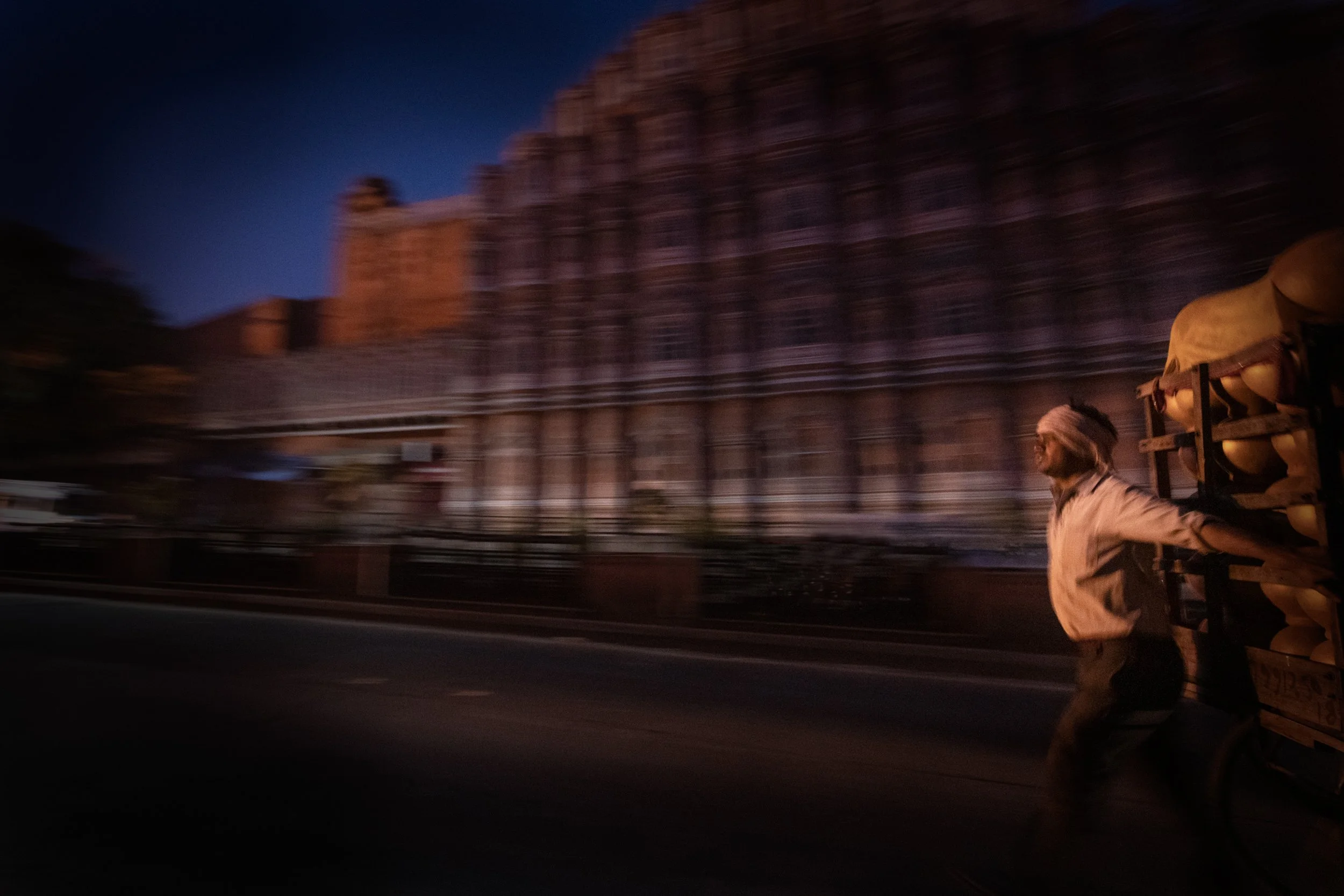 Motion-blur street scene of a man pushing a loaded cart past illuminated palace architecture at dusk in Udaipur, India.
