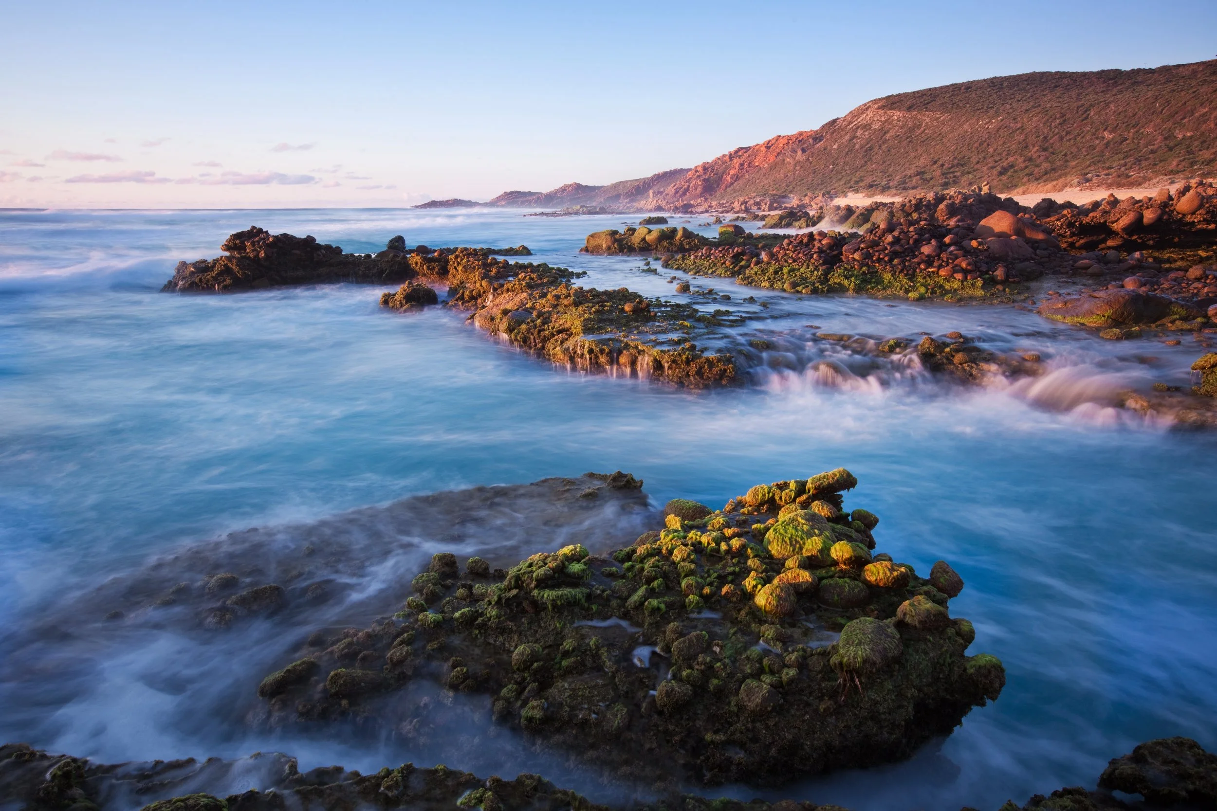 Rocky coastline at Injidup Beach near Margaret River, Western Australia, with blue ocean, moss-covered rocks, and warm sunset light.