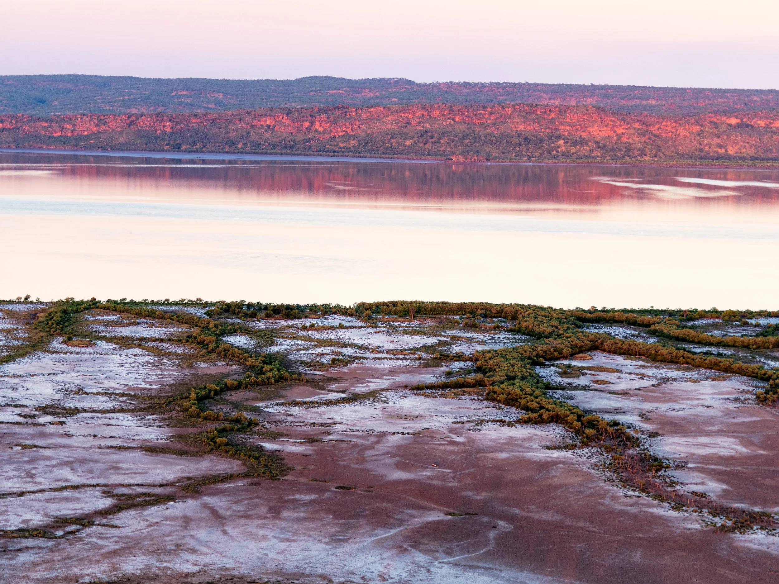 Pink and silver Kimberley tidal flats at sunset with still water, mangroves, and red escarpment reflections.