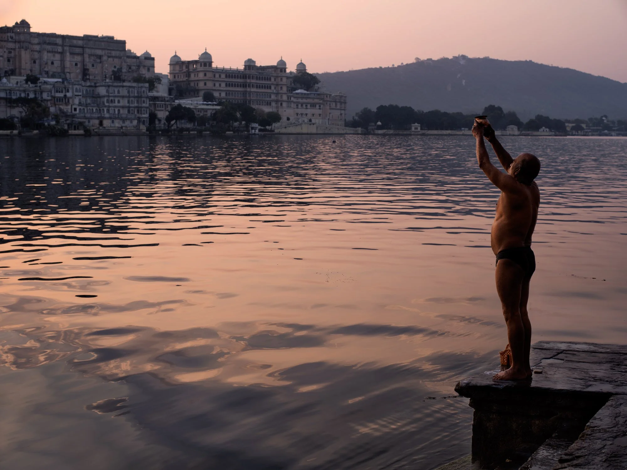 Silhouetted man performs a prayer ritual beside Lake Pichola at sunset in Udaipur, with rippling water and palace reflections.