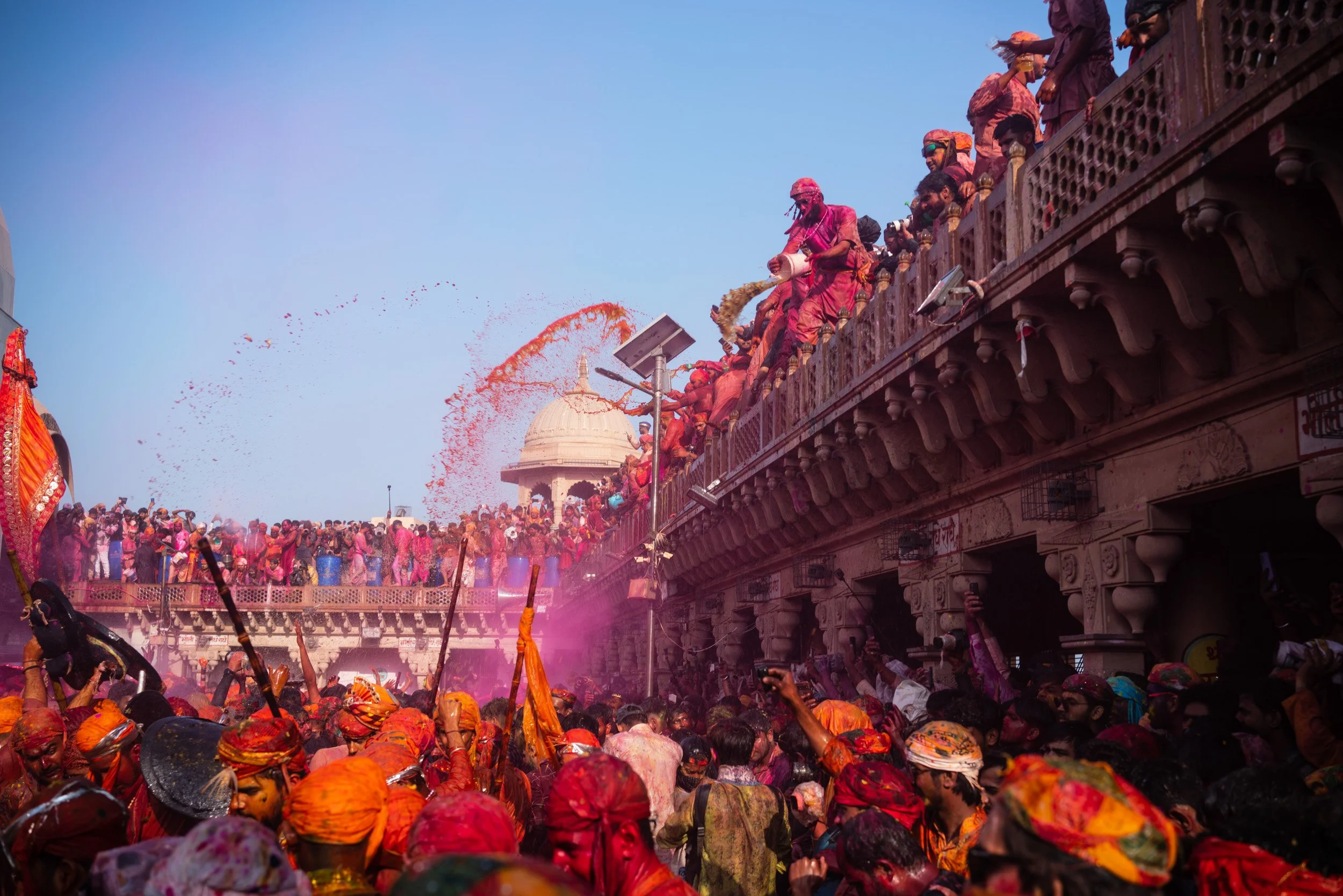 Crowds celebrate Holi at a Hindu temple courtyard as people on balconies throw bright red and pink gulal powder over festival participants below.