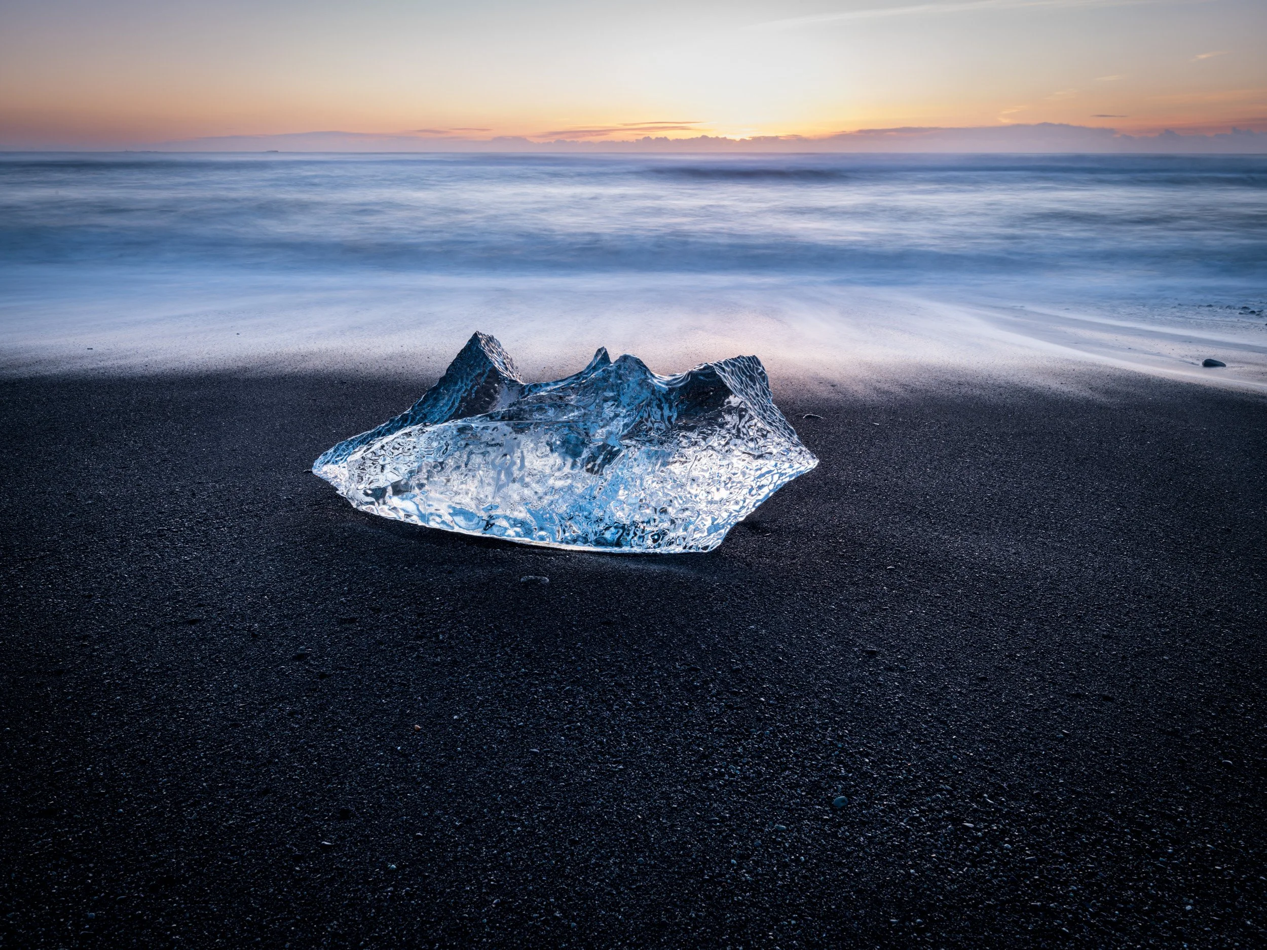 Blue iceberg resting on black volcanic sand at Iceland’s Diamond Beach, with soft ocean waves and warm sunrise light.