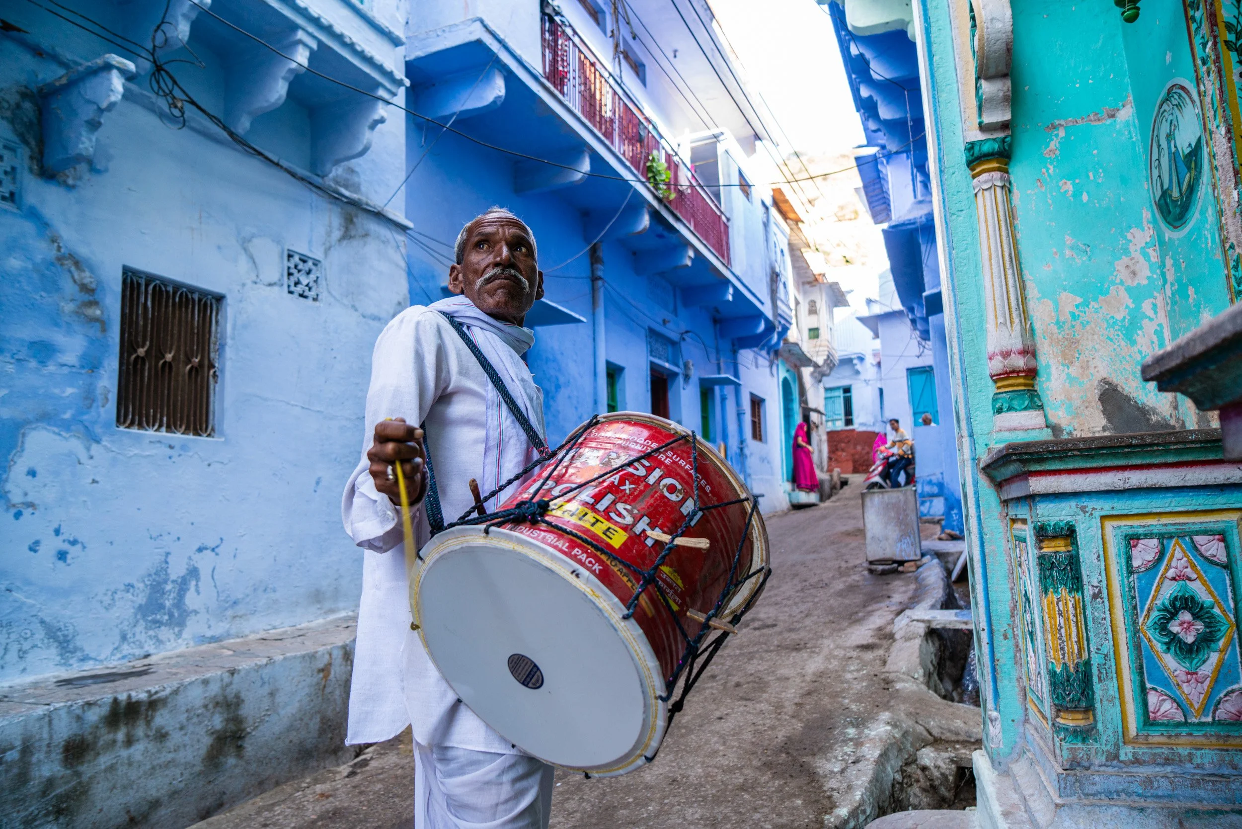 Elderly drummer in white clothing carries a large red dhol through a narrow blue-painted Indian street in Bundi, India.
