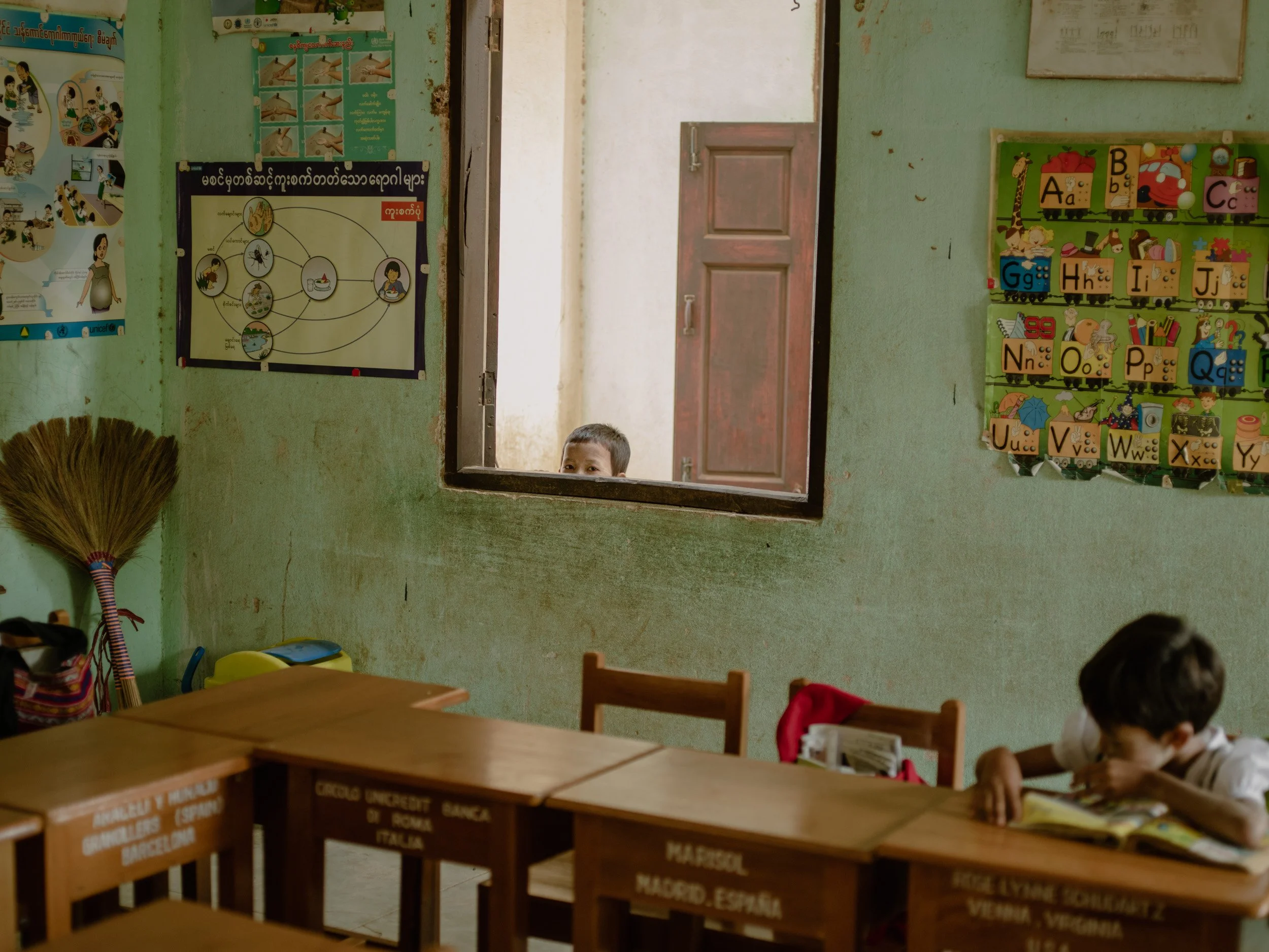 Quiet Myanmar classroom interior with alphabet posters, wooden desks, and a child peeking through an open window.