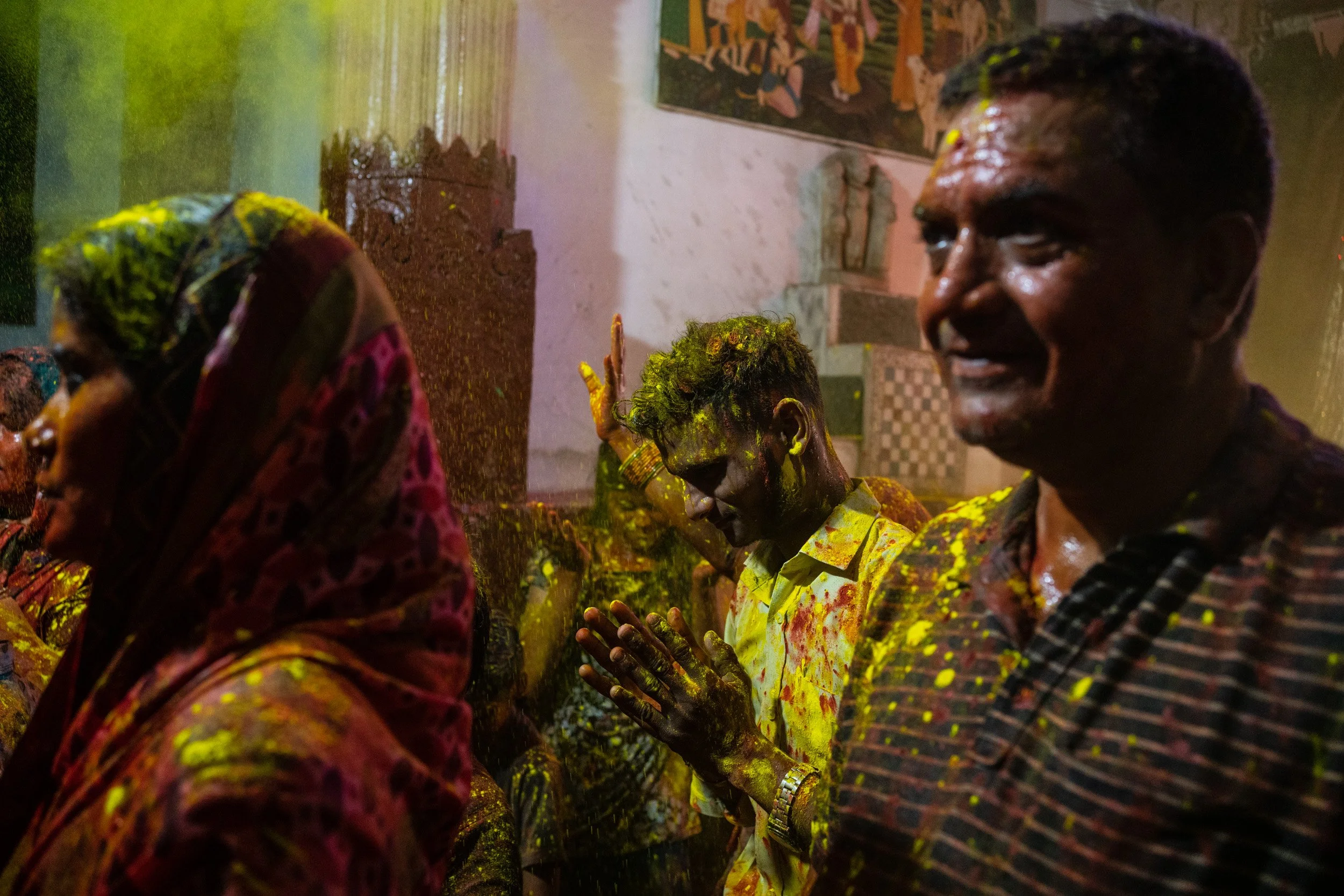 Devotees covered in yellow Holi powder pray inside a temple, with smiling worshippers surrounded by bright gulal and festive color.