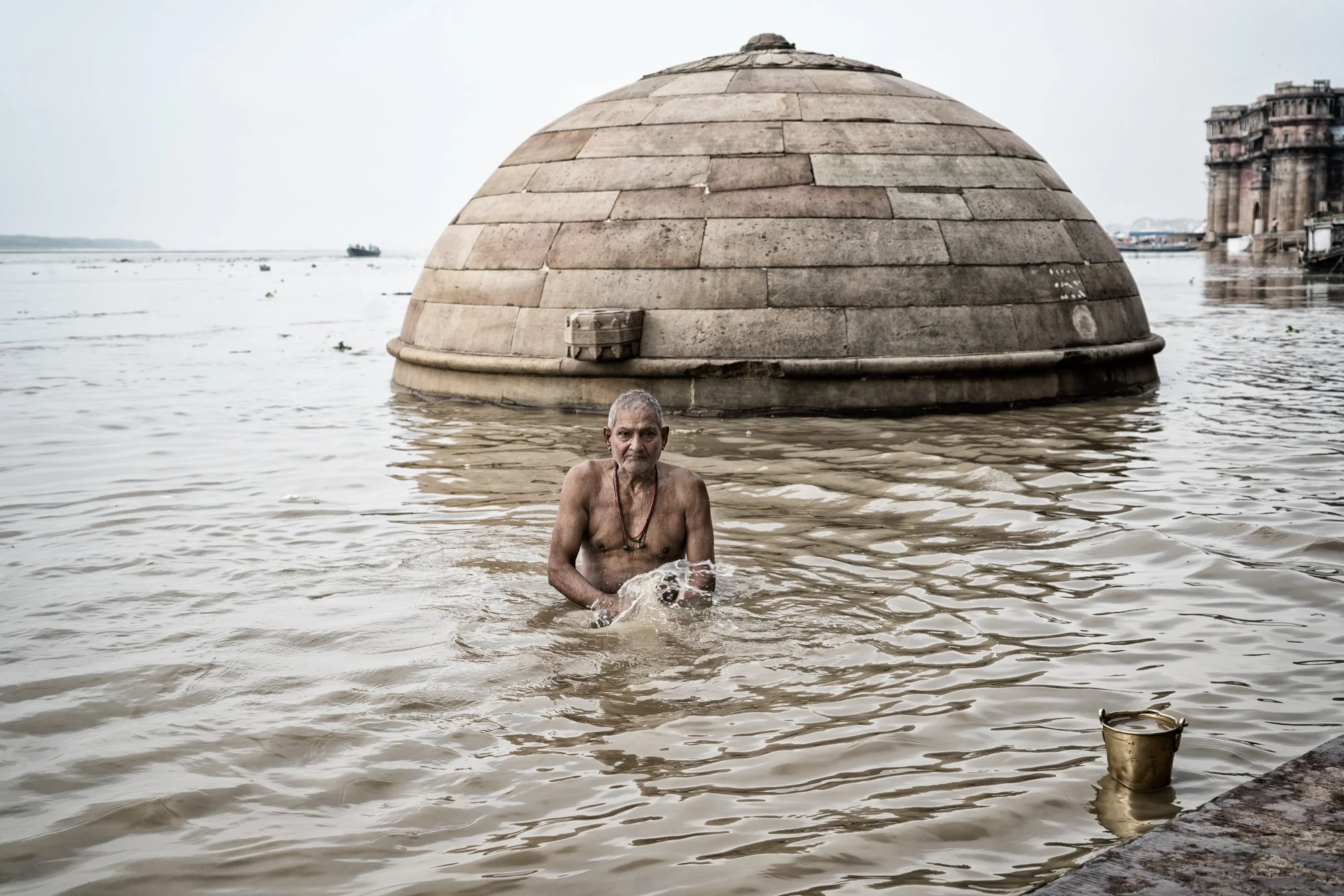 Elderly man bathes in the Ganges River near a stone temple dome at Varanasi ghats, India.