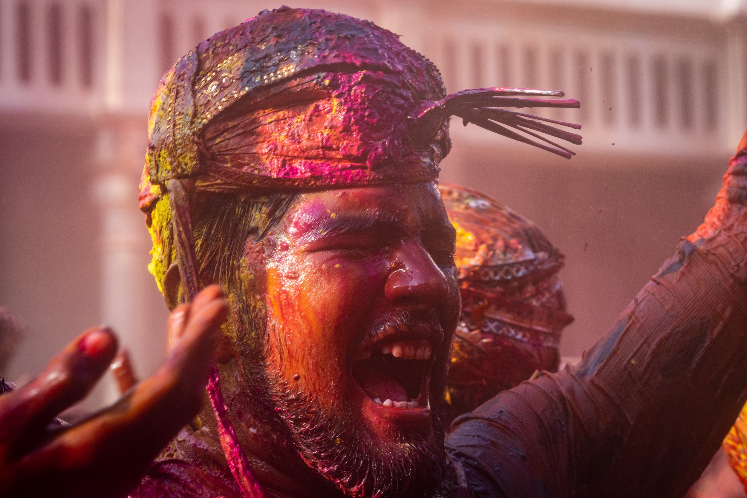 Joyful man with a colorful turban and face covered in pink, yellow, and red gulal celebrates Holi in a crowded festival scene.