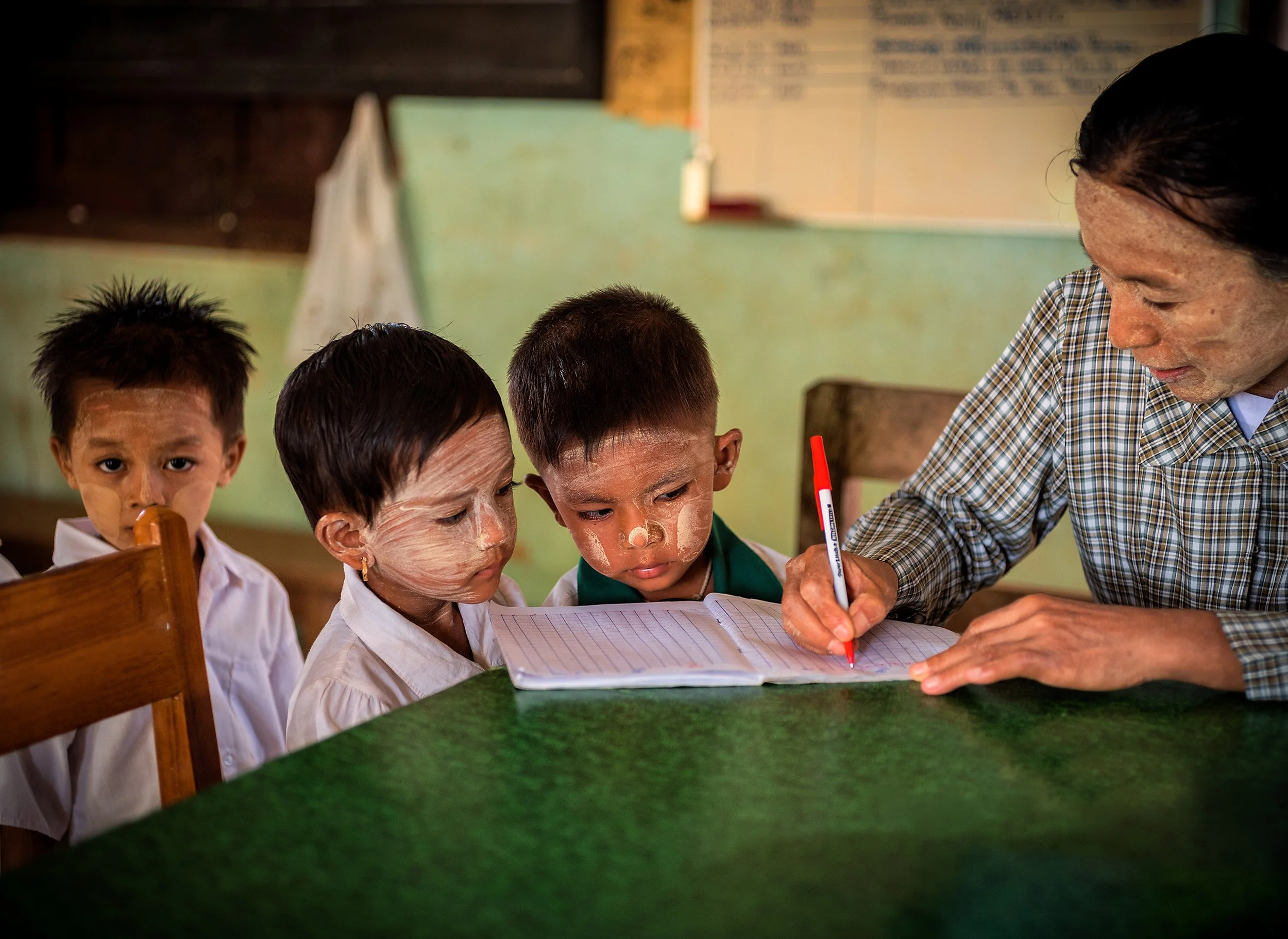 Teacher writes in a notebook while young schoolchildren with traditional face paint gather around a classroom desk.