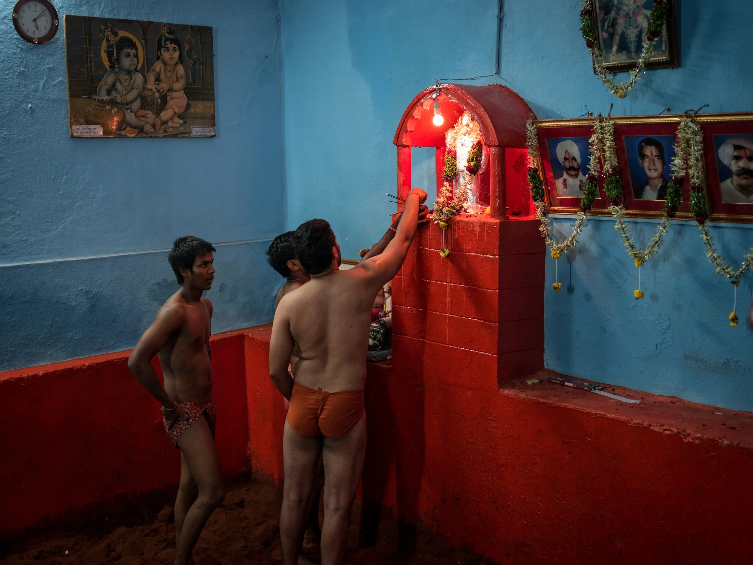 Three traditional Indian wrestlers in loincloths perform a ritual at a glowing red shrine inside a blue and red mud-floor akhara, with framed religious images and garlands on the wall