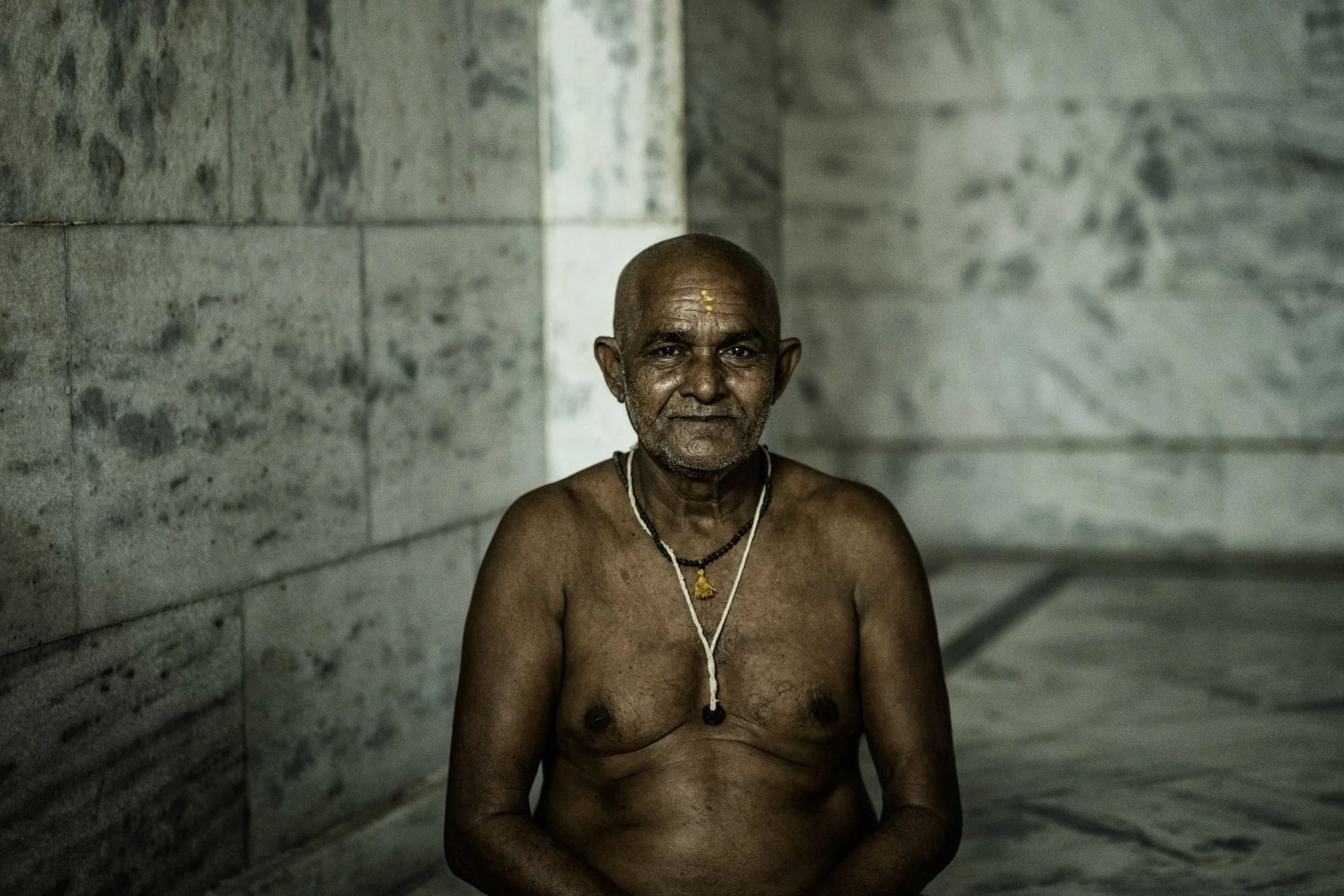 Elderly shirtless holy man with a necklace sits calmly against a marble wall in a quiet spiritual portrait.