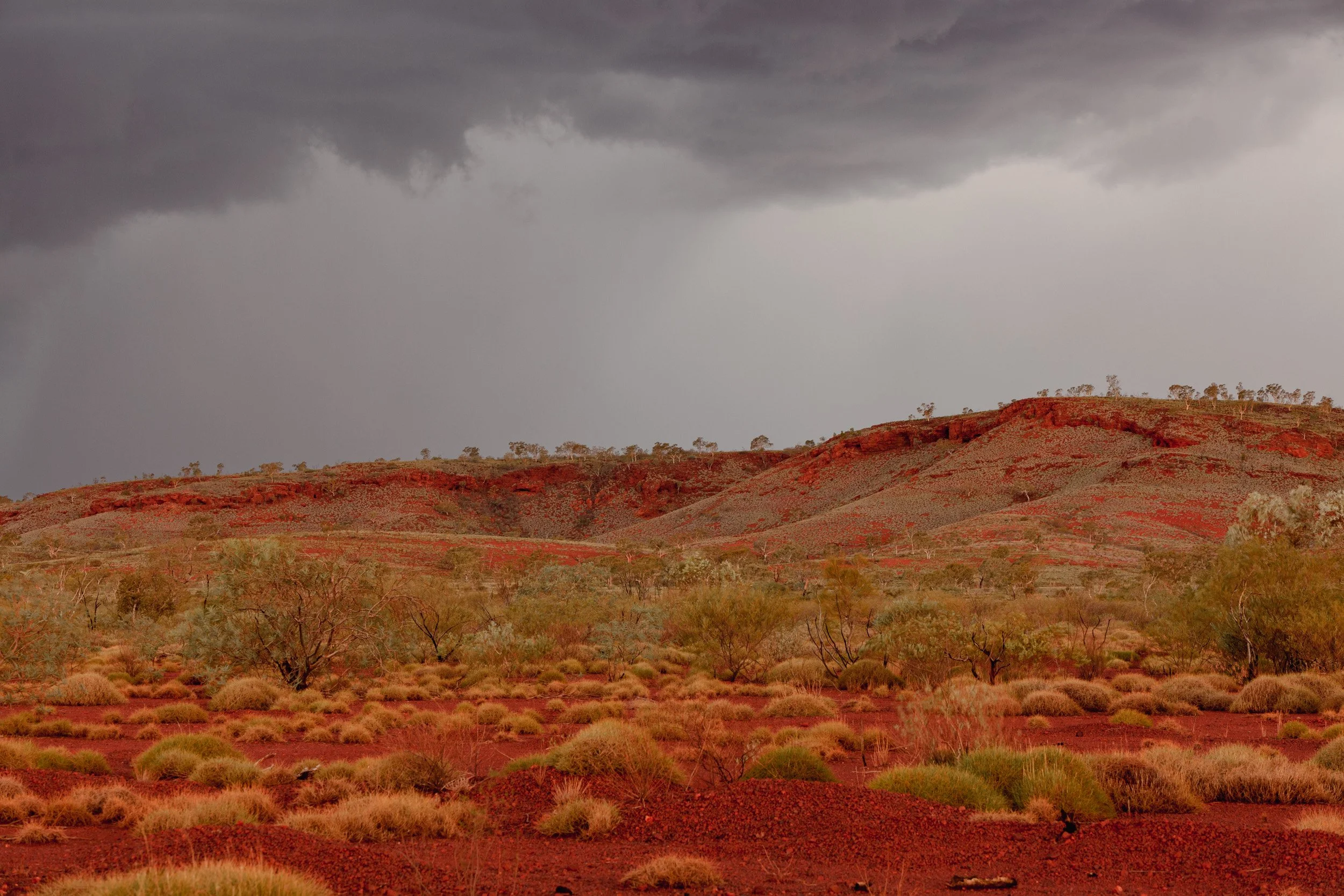 Storm clouds gather over a red Pilbara outback landscape with low hills and desert scrub.