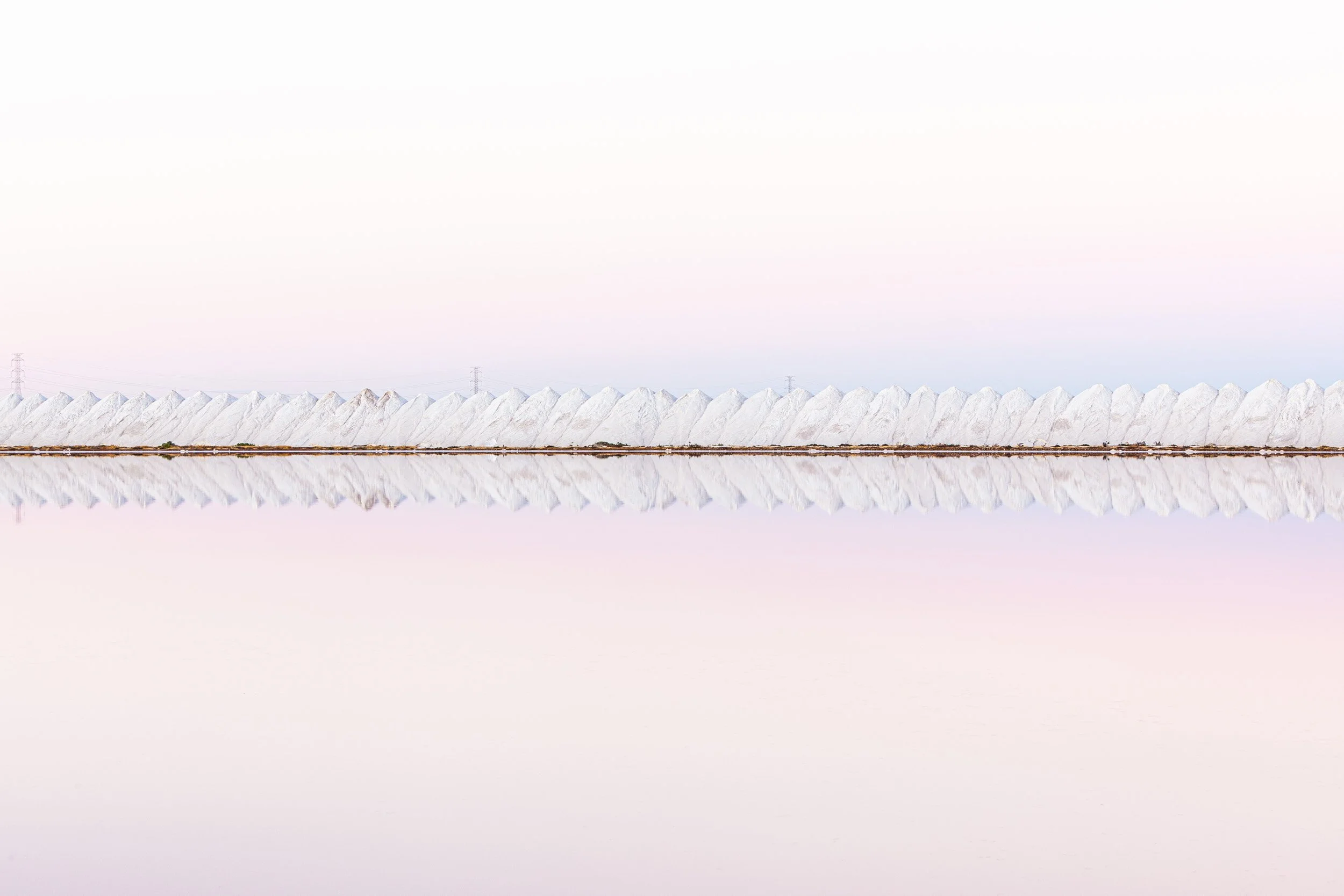 Soft pastel minimalist landscape of salt piles with a thin horizon line, calm water, and distant shoreline in muted pink and white tones.