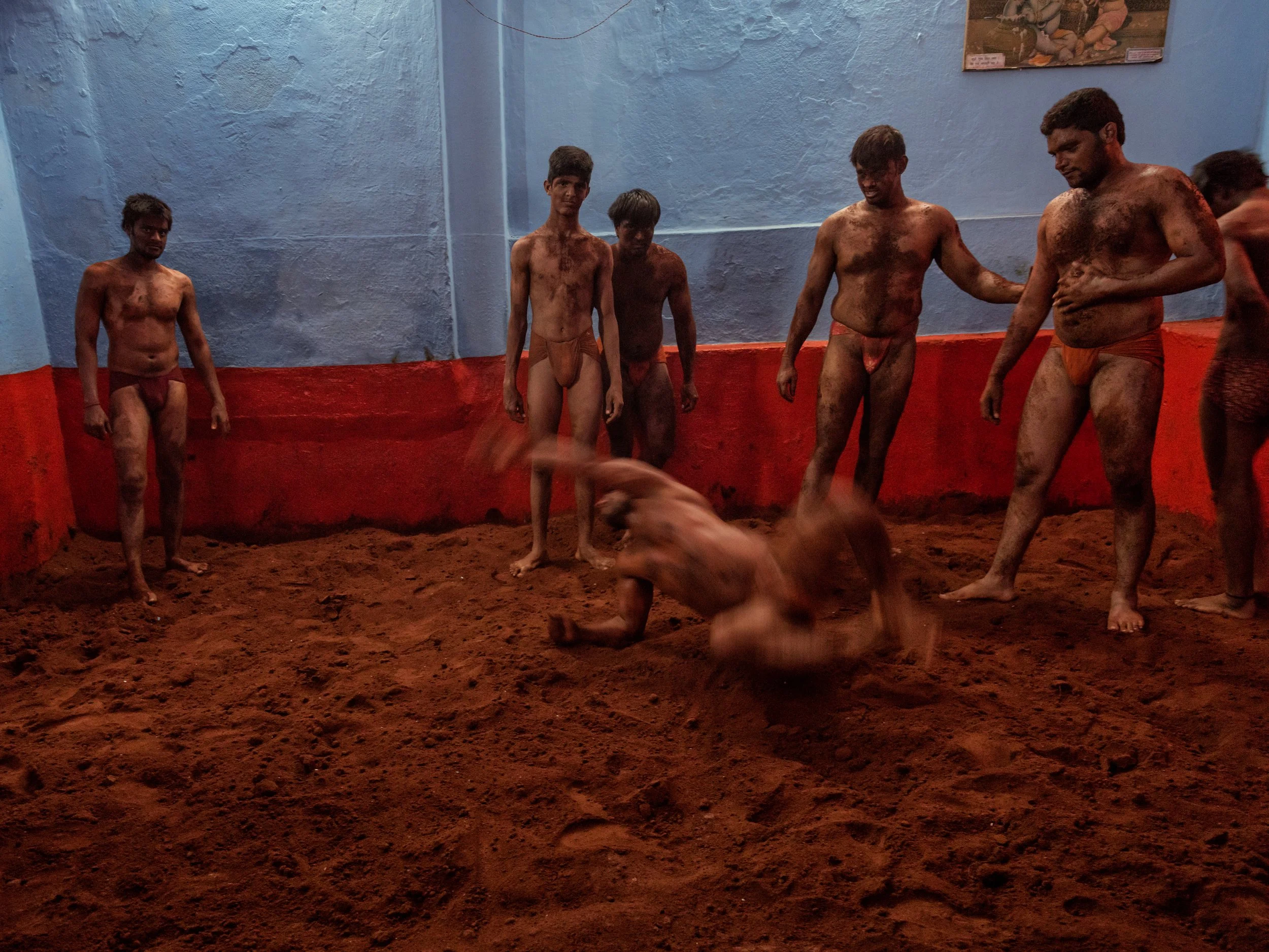 Mud-covered Indian kushti wrestlers practice a fast takedown in a red clay akhara pit while fellow wrestlers watch against blue and red training-room walls.