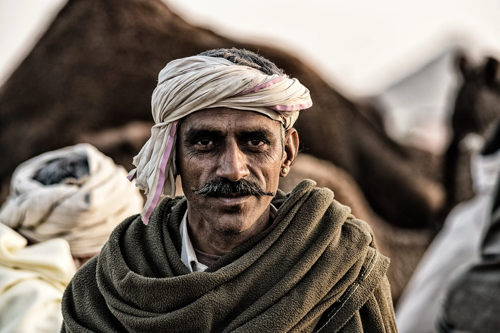 Indian camel trader with a white turban, curled moustache, and wool shawl poses outdoors in a dramatic rural portrait.