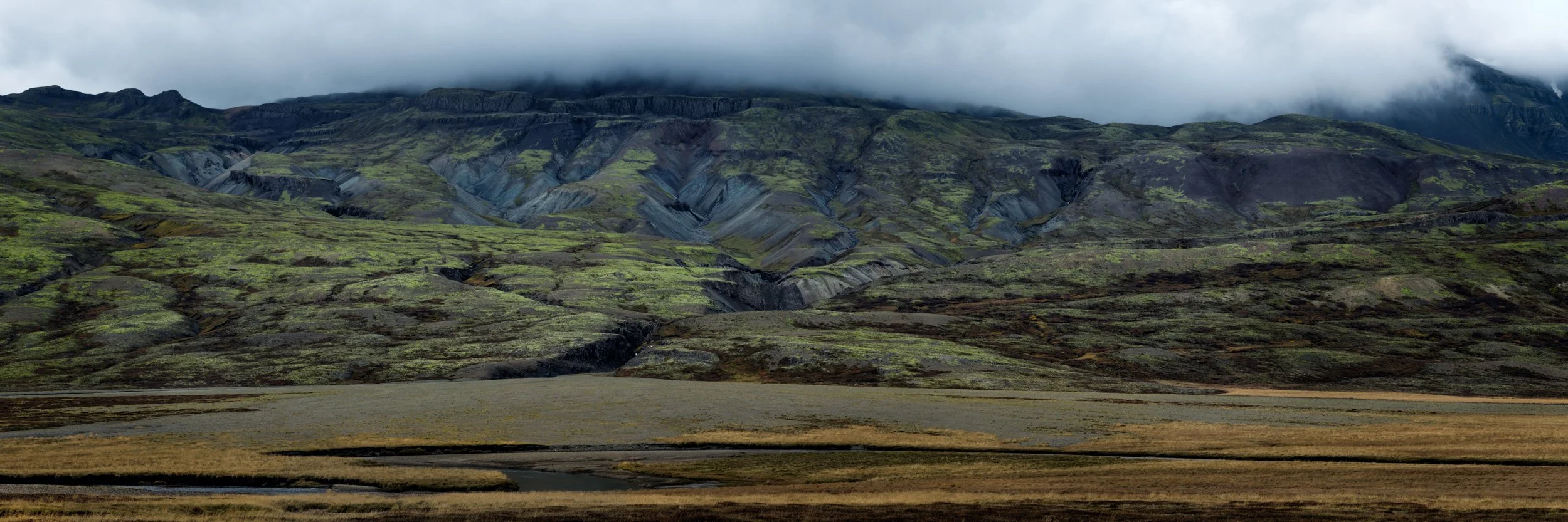 Wide Icelandic mountain and lava field landscape under low clouds, showing green mossy terrain and dramatic volcanic ridges.