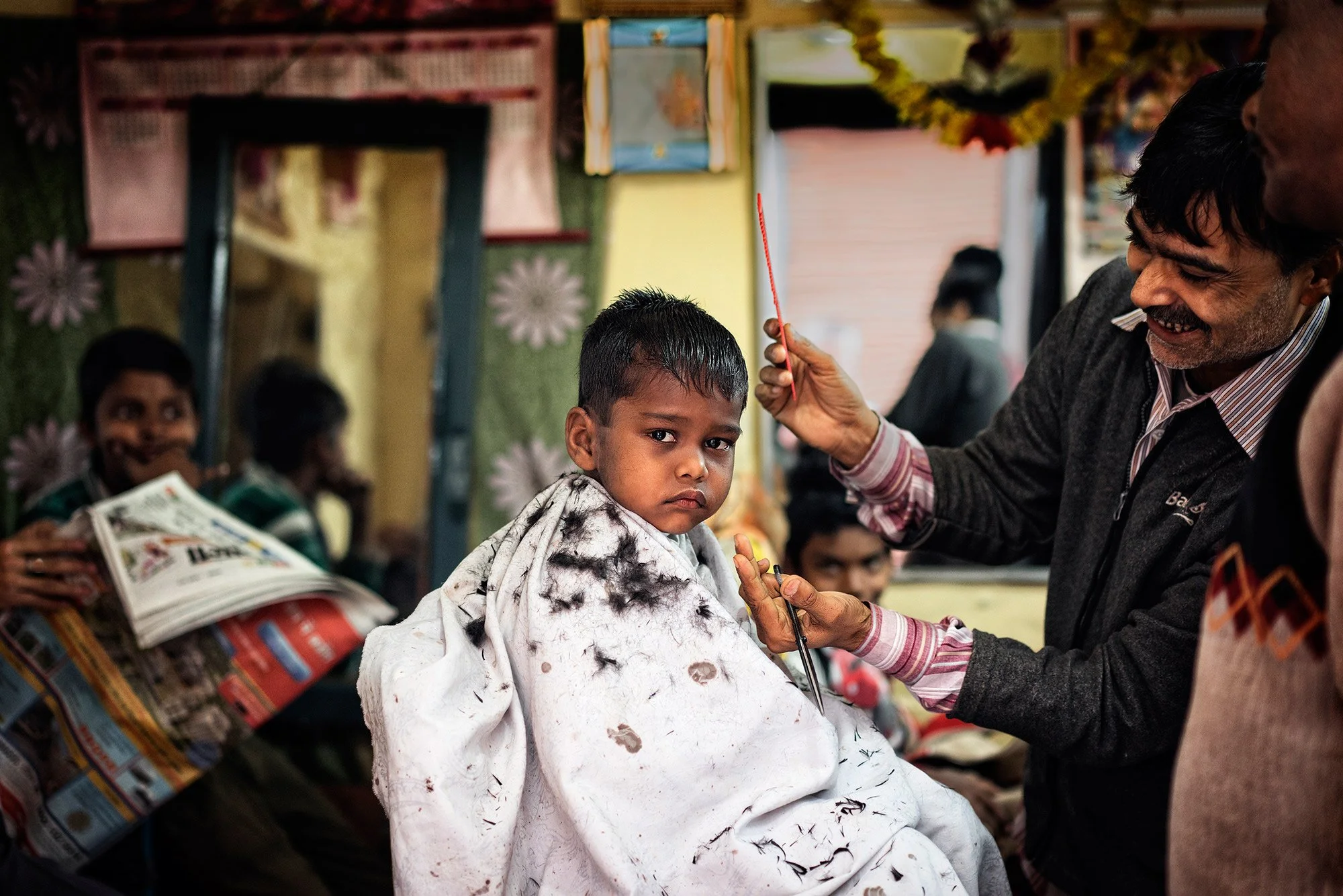 Barber smiles while cutting a young boy’s hair inside a small local barbershop, with the child wrapped in a white barber cape.