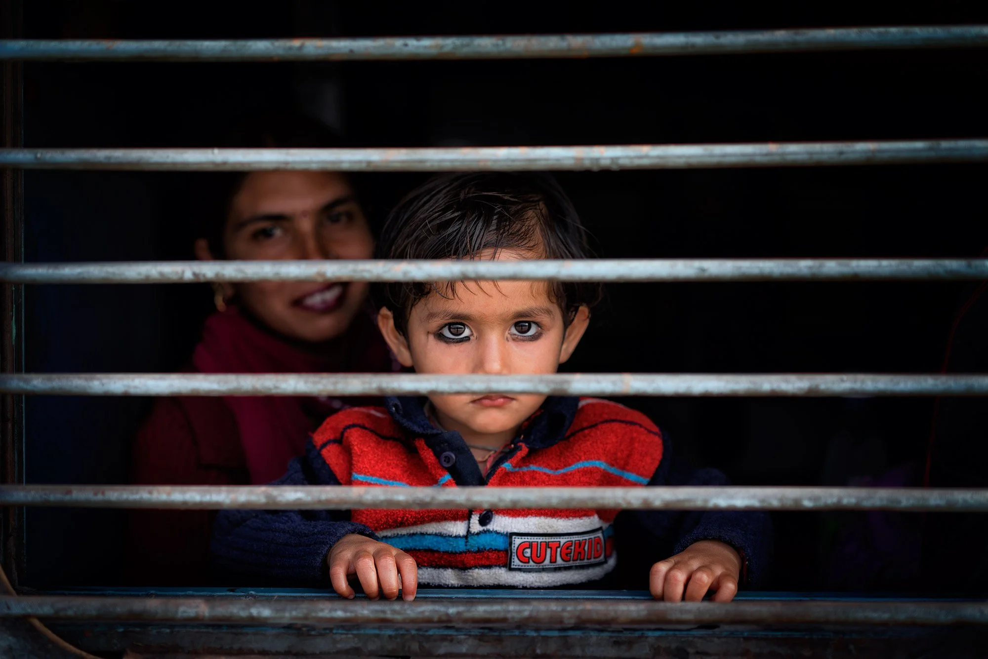 Young child in a red sweater looks through horizontal metal bars, with a woman seated in shadow behind him during a candid travel portrait.