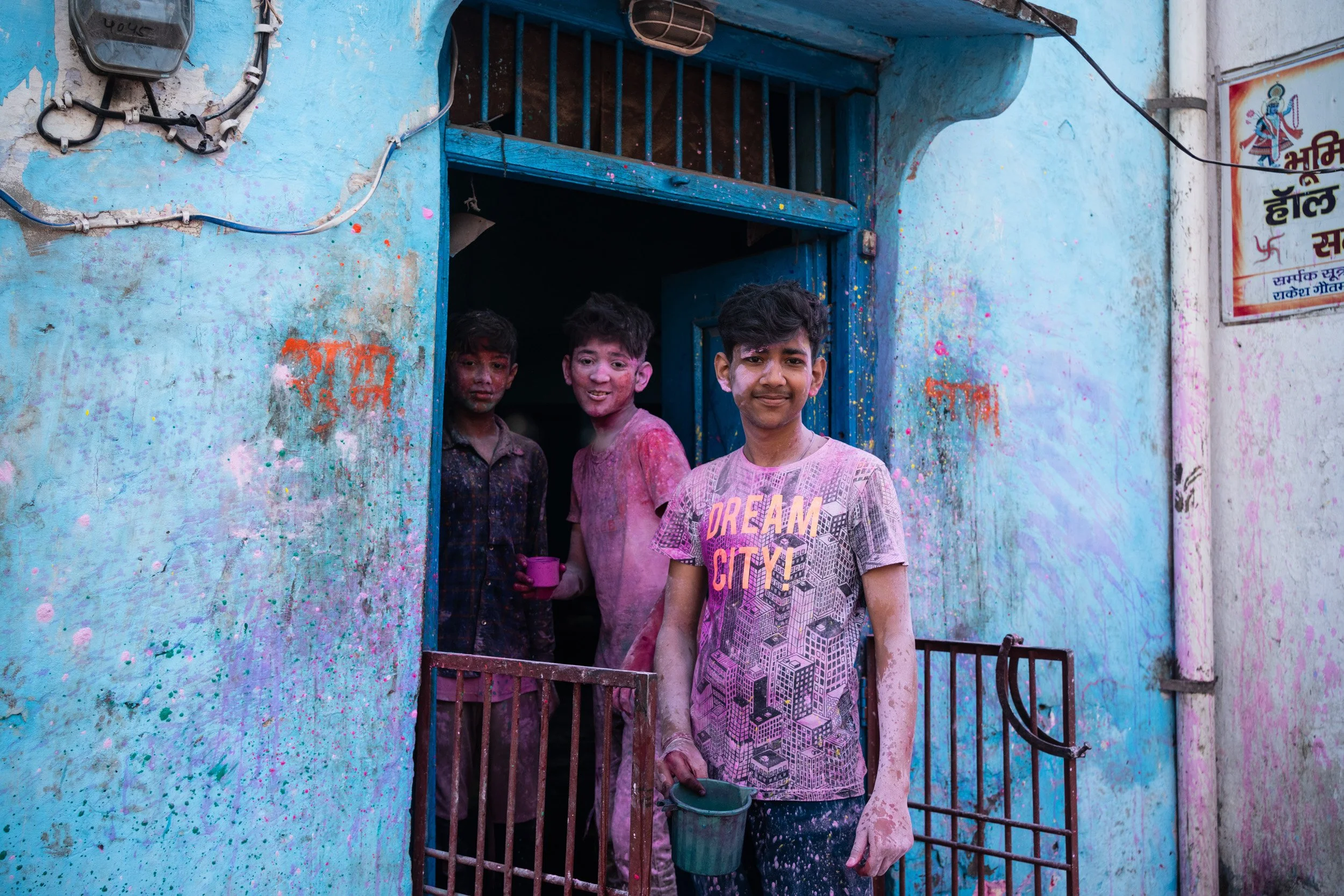 Three boys covered in pink and purple Holi powder stand in a blue doorway holding small buckets during a neighborhood color festival.