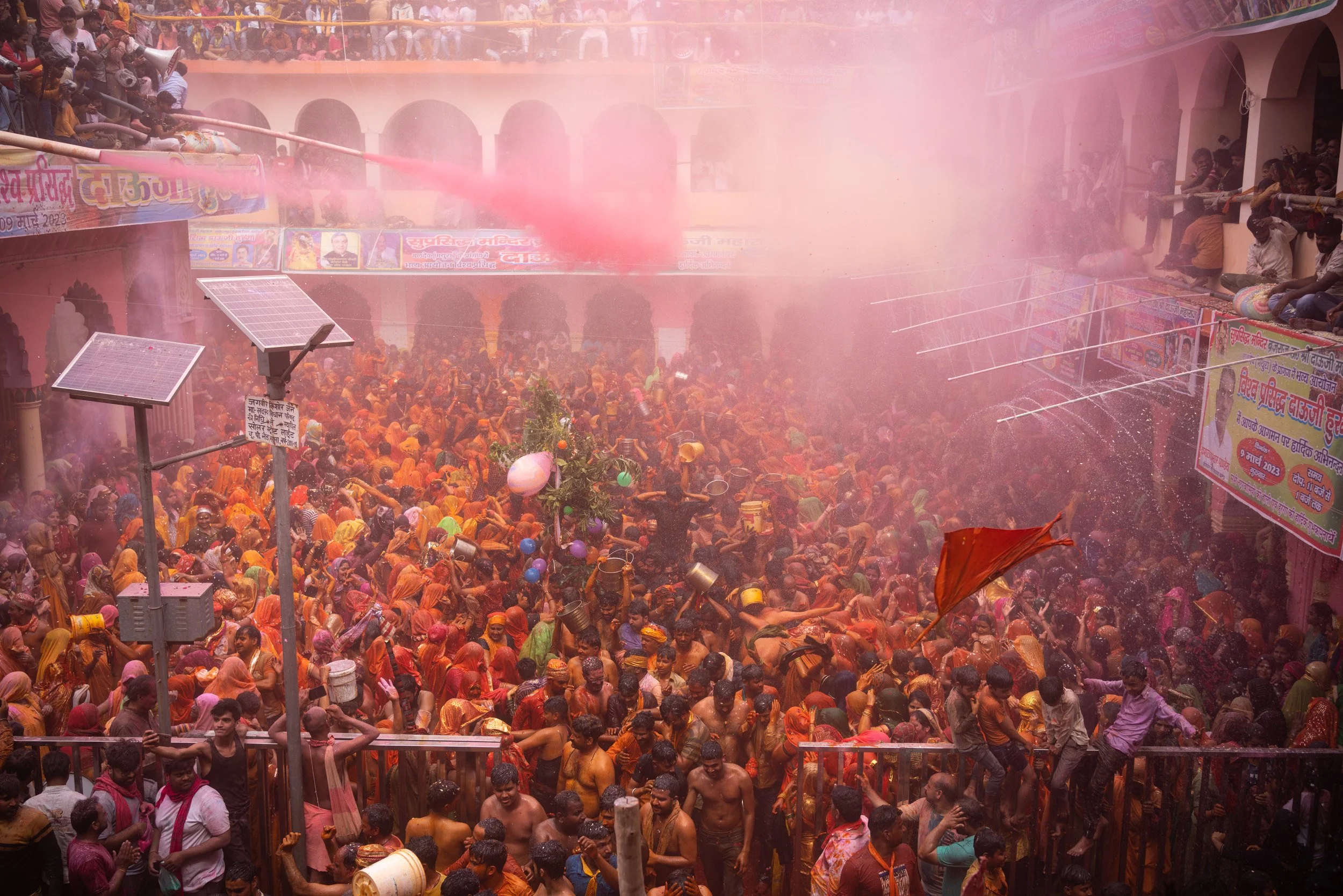 Massive Holi celebration in a temple courtyard, with dense crowds in orange clothing, flying water spray, pink powder haze, and raised buckets.