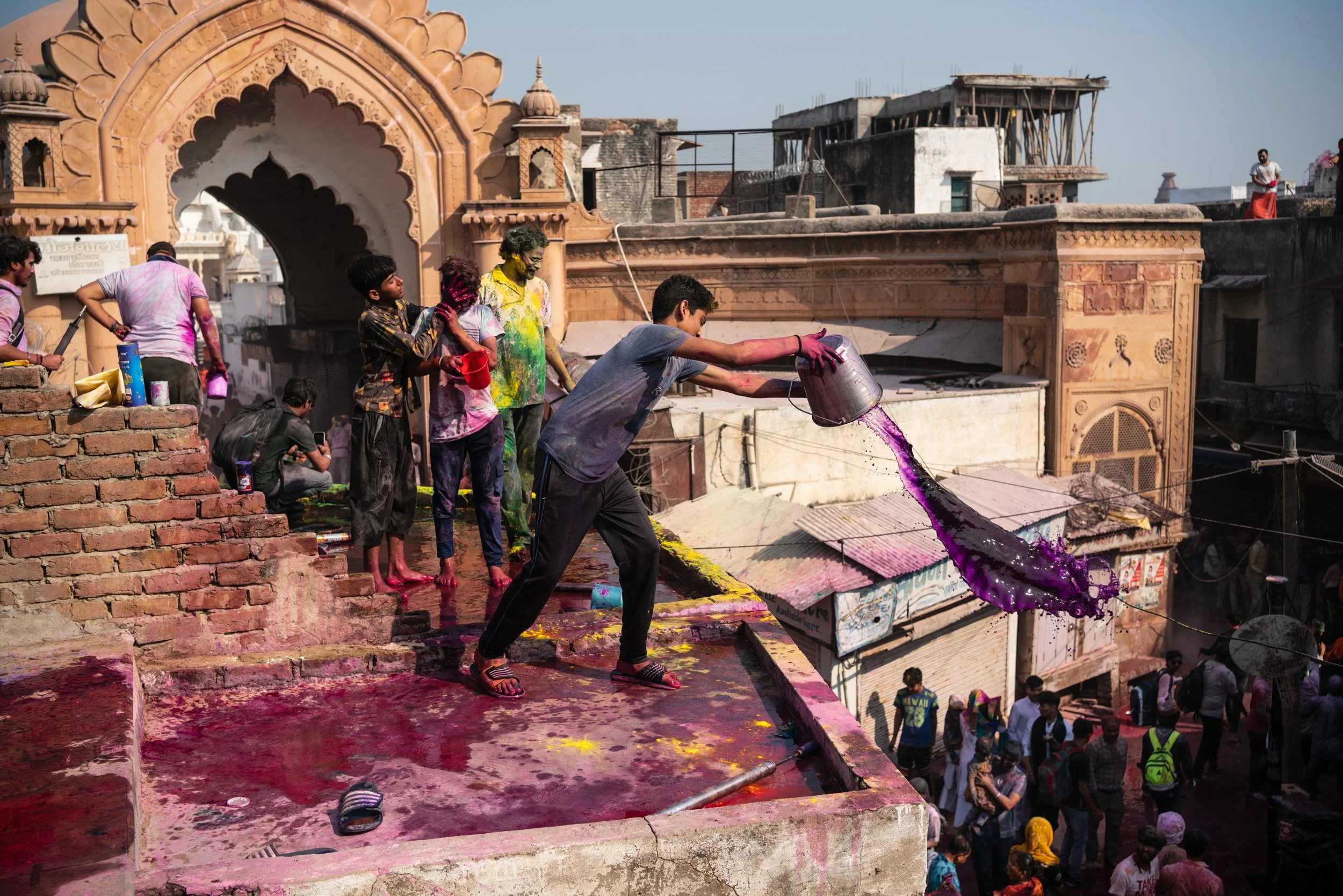 Boy pours purple Holi water from a metal bucket off a rooftop while friends covered in festival colors stand near a temple entrance.
